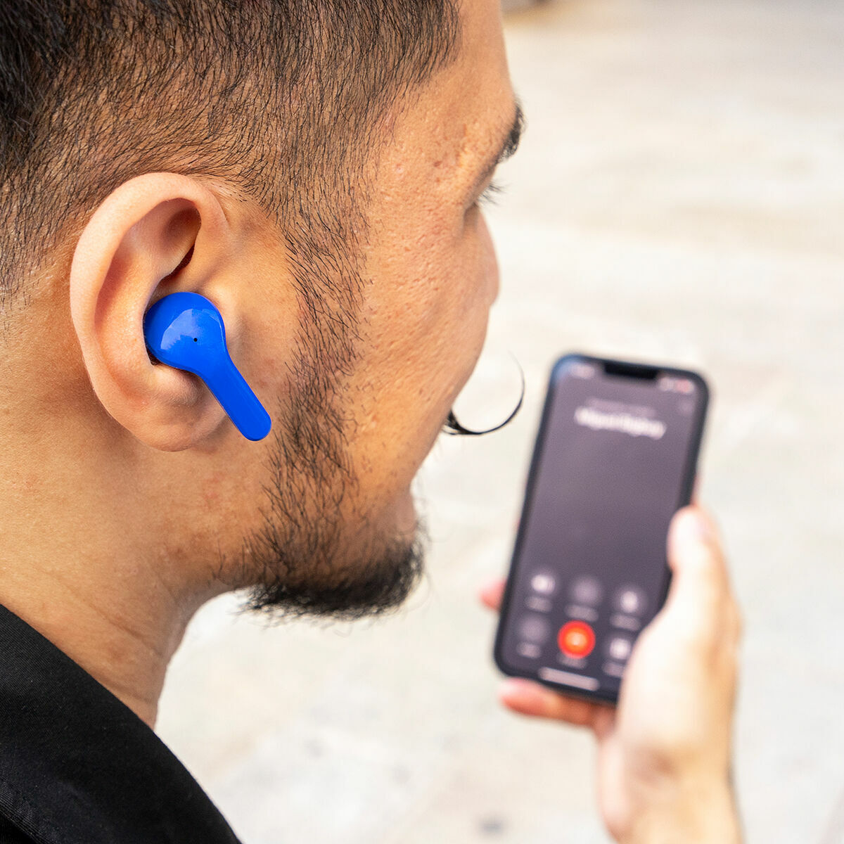 Mann mit blauem Ohrhörer im Ohr, hält ein Telefon mit Anrufbildschirm. Nahaufnahme, unscharfer Hintergrund.