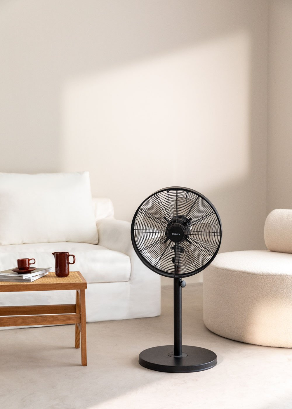Ventilador eléctrico moderno sobre un soporte. Un sofá blanco y una mesa pequeña están cerca.