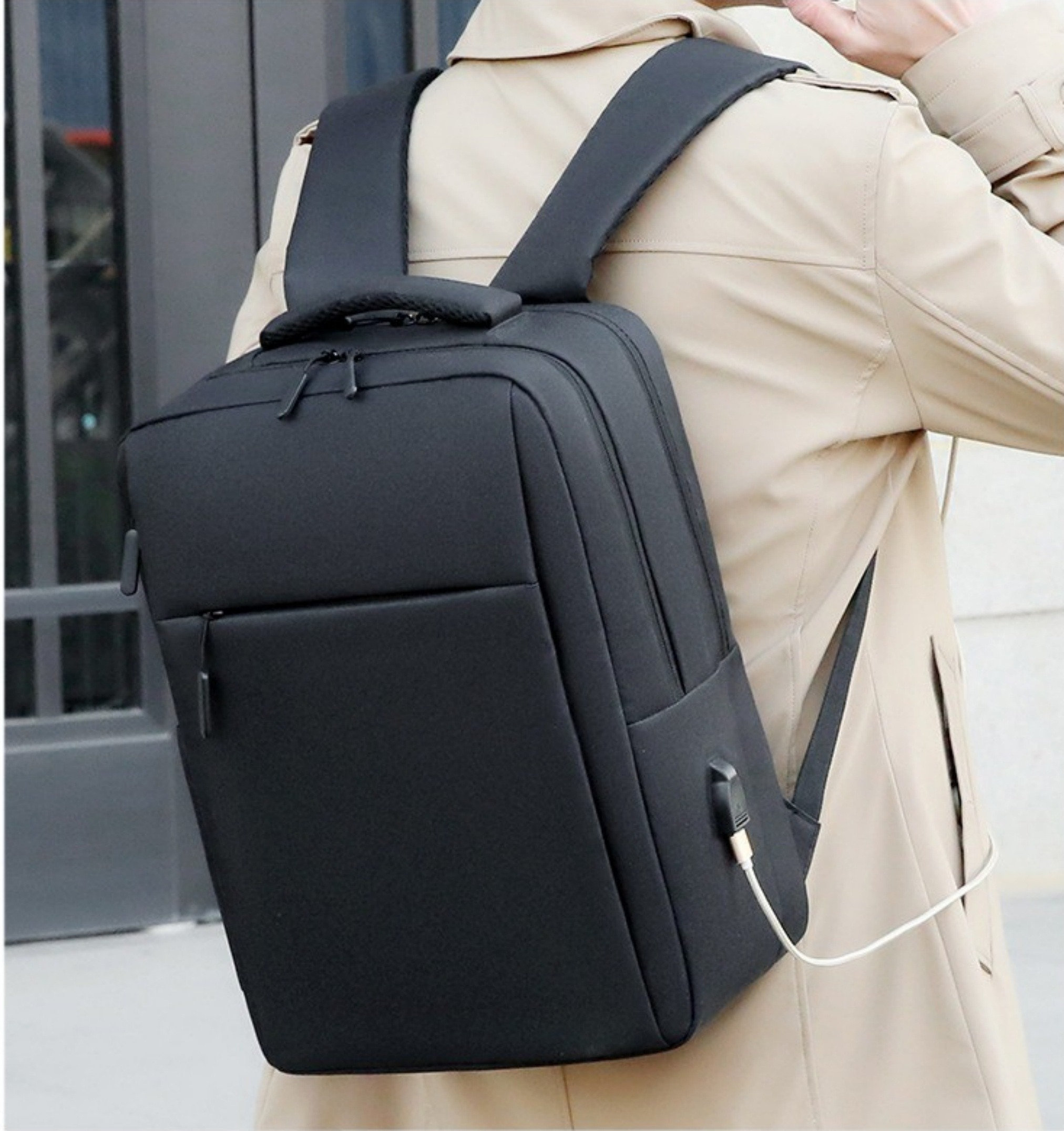 Ein schwarzer Rucksack auf dem Rücken einer Person. Die Person trägt einen beigefarbenen Mantel. Der Rucksack hat einen USB-Anschluss.