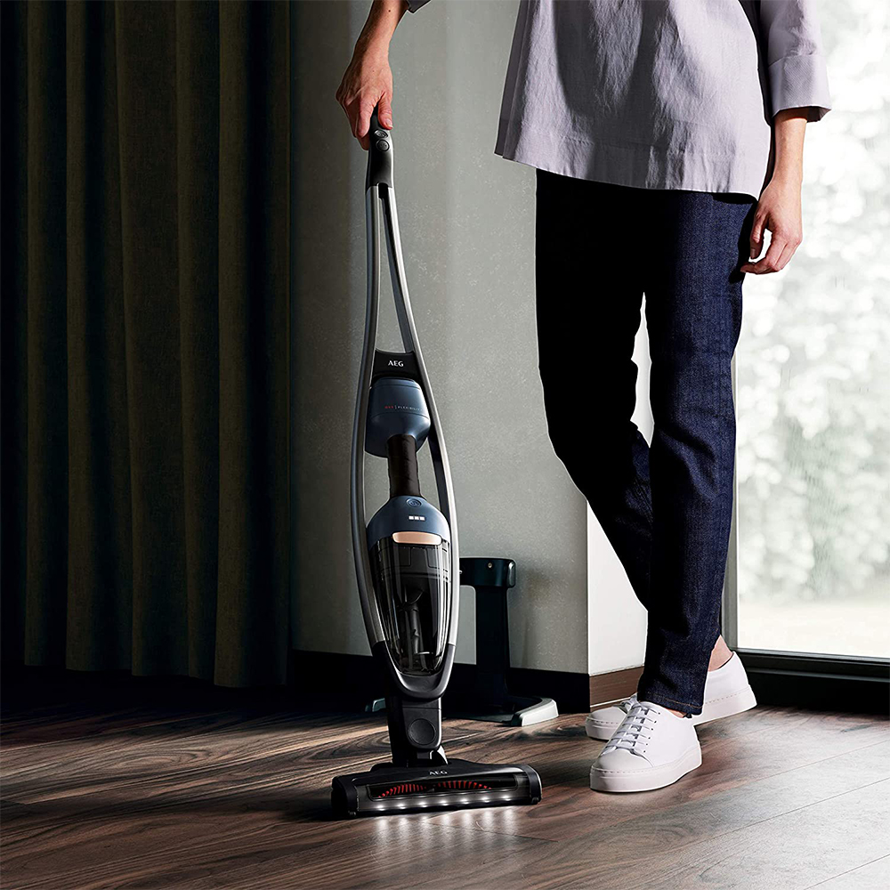 Eine Person hält einen grau-schwarzen Staubsauger auf einem Holzboden. Die Person trägt dunkle Jeans.