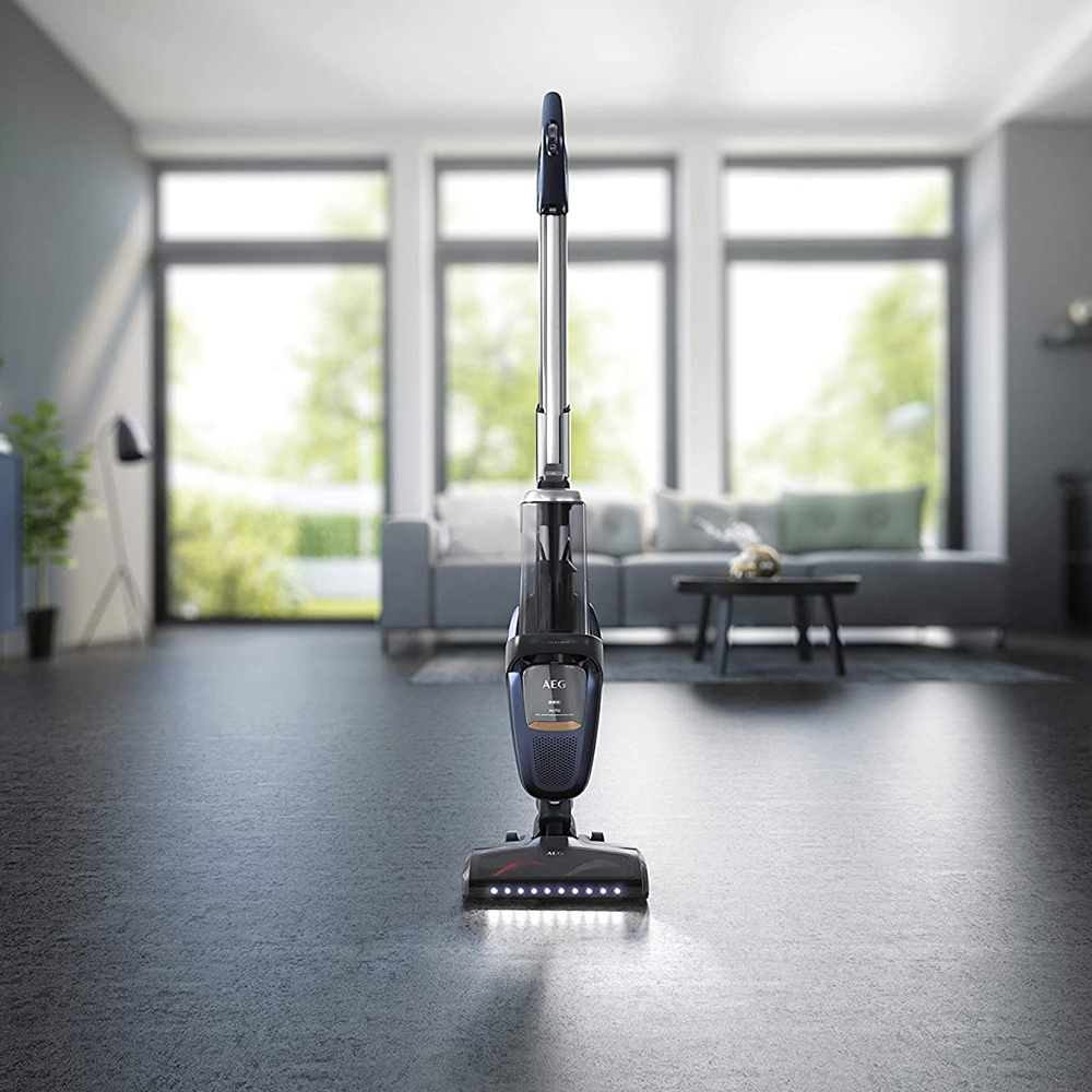 Ein kabelloser Staubsauger steht auf grauem Boden. Der Hintergrund ist ein Wohnzimmer.