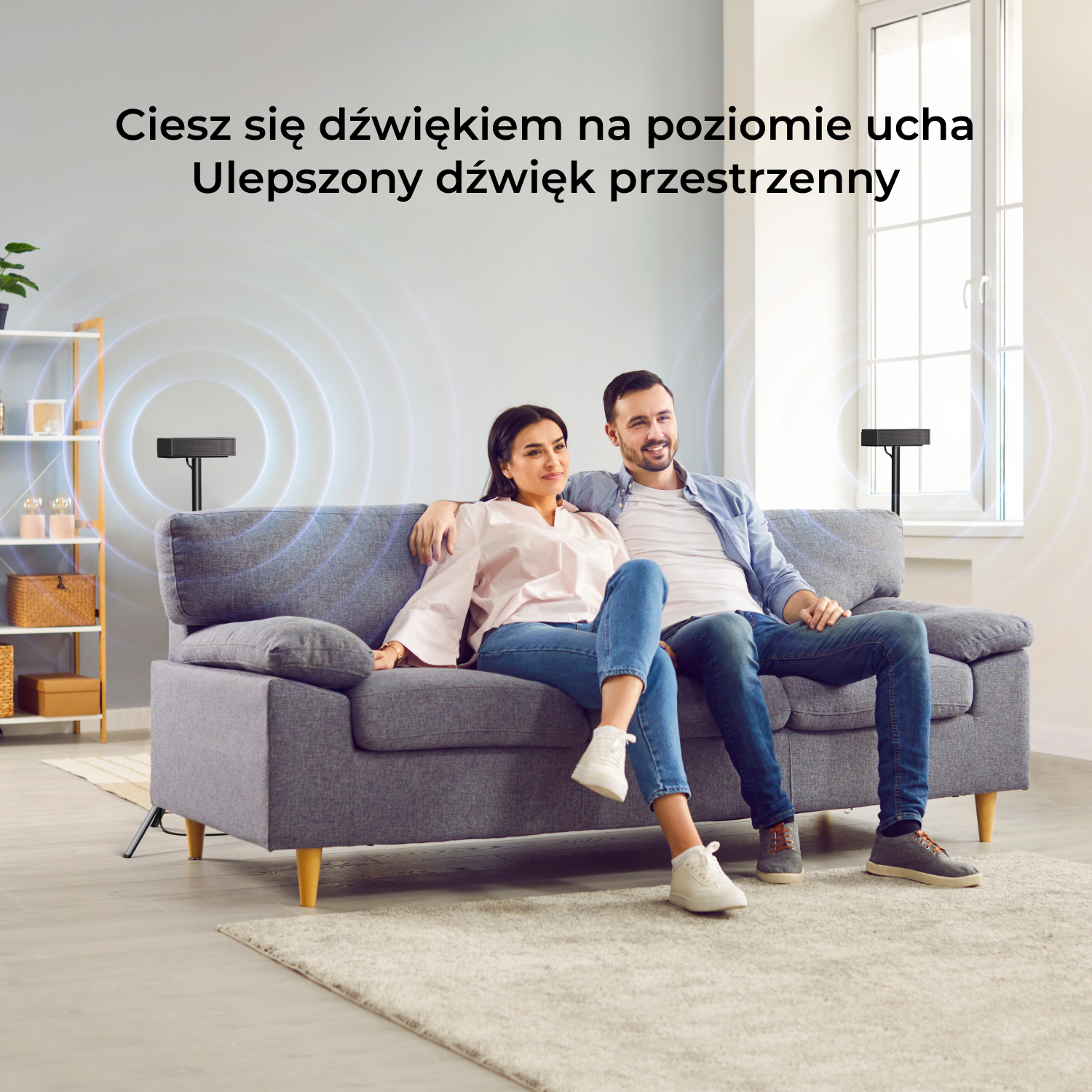 Para siedzi na szarej sofie, z głośnikami na stojakach po obu stronach.
