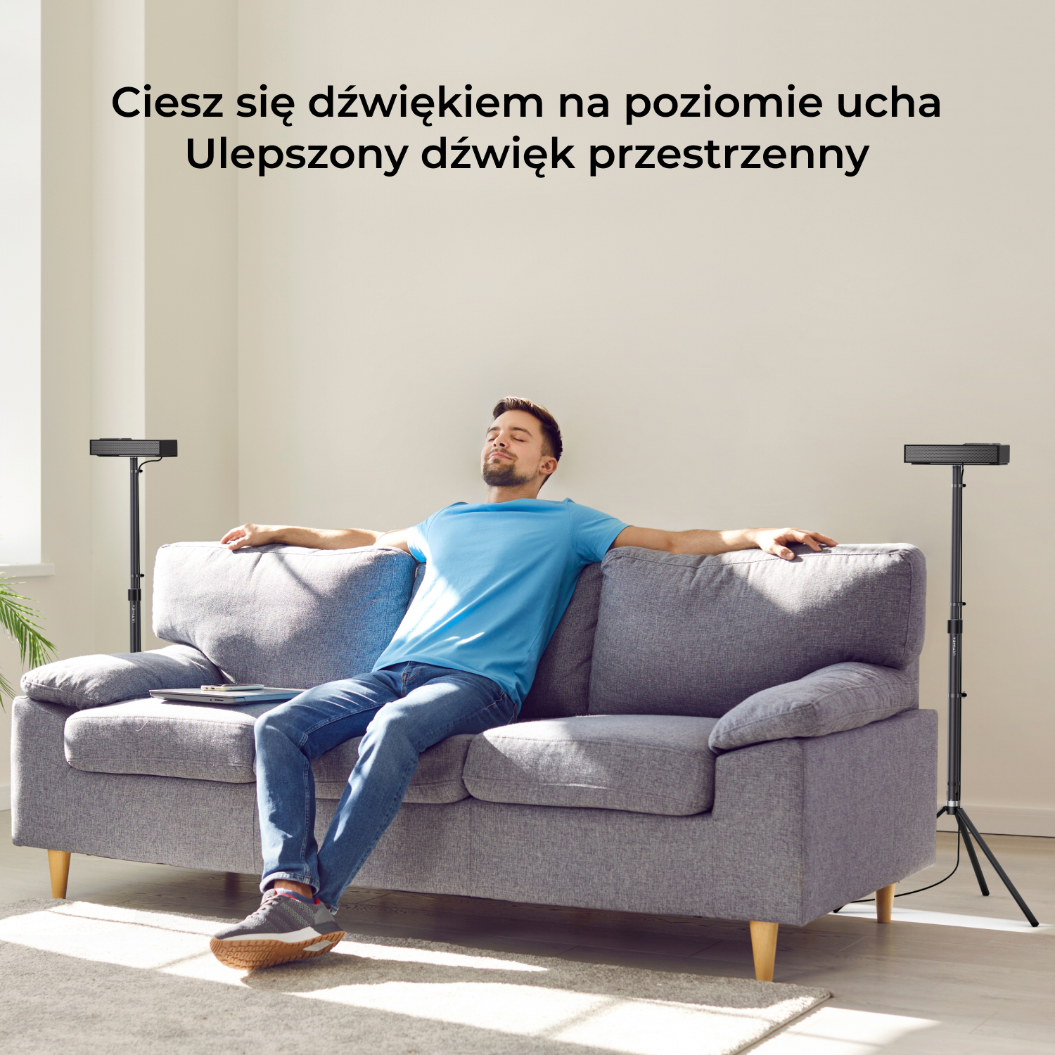 Mężczyzna zrelaksowany na szarej sofie z dwoma głośnikami na stojakach. Ustawienie w salonie.