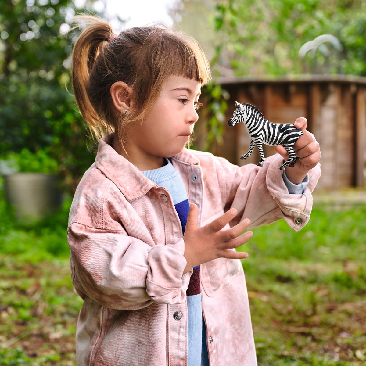 Jeune fille tenant un jouet zèbre, portant une veste rose, dans un jardin.