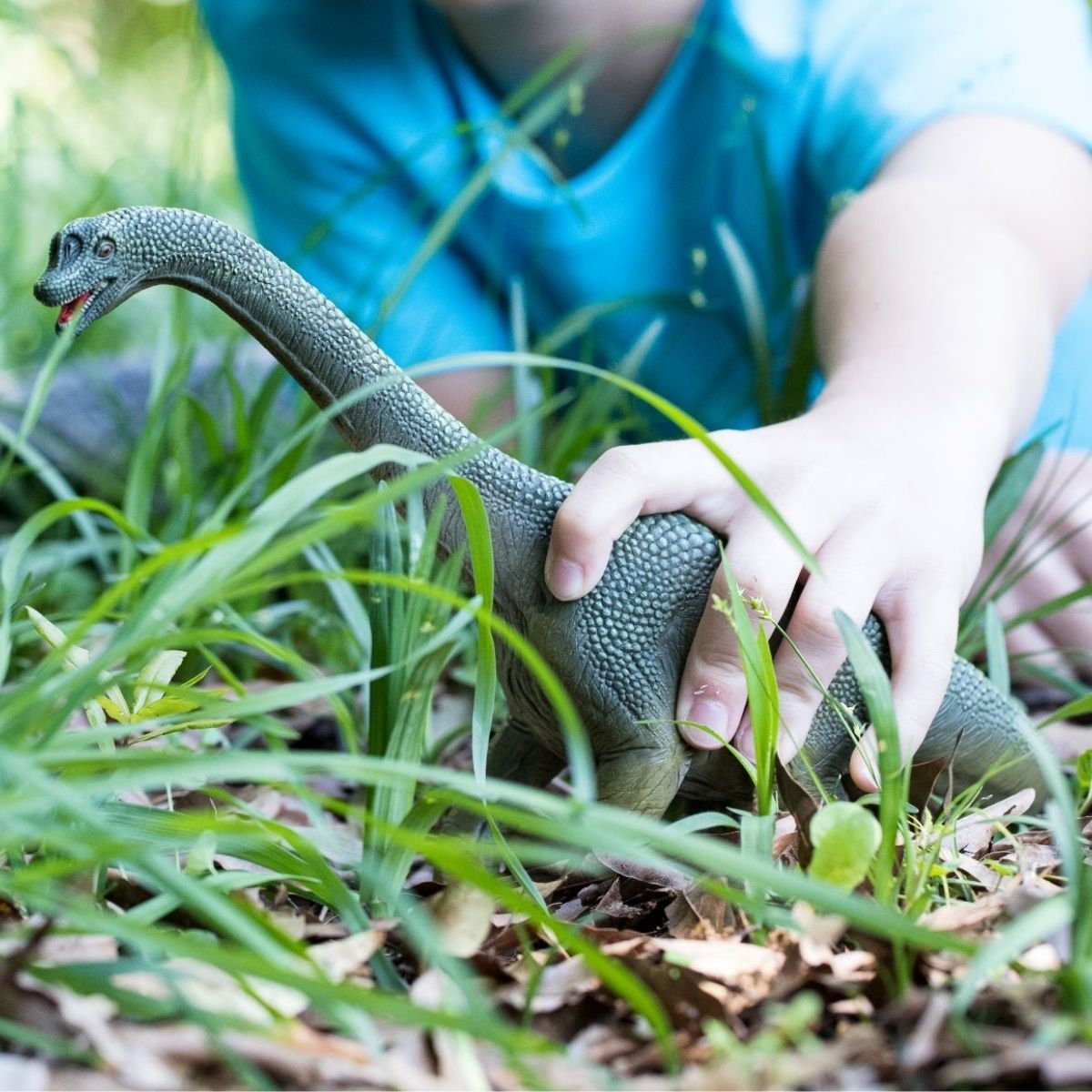 Kinderhand hält ein grünes Dinosaurier-Spielzeug in hohem Gras.