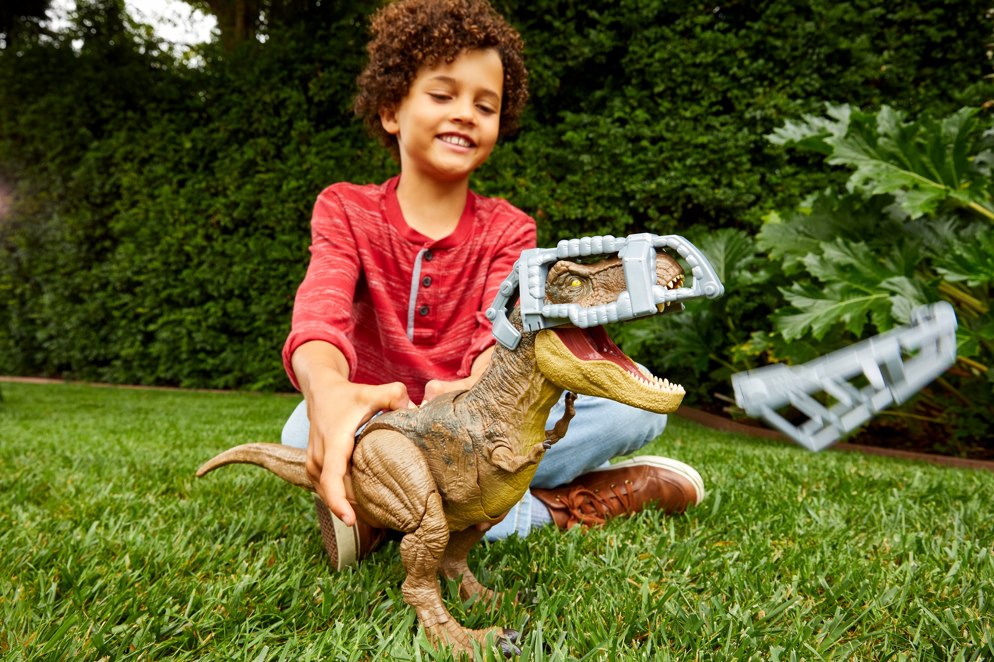 Niño con un dinosaurio de juguete en las manos. Hierba verde y un seto en el fondo.