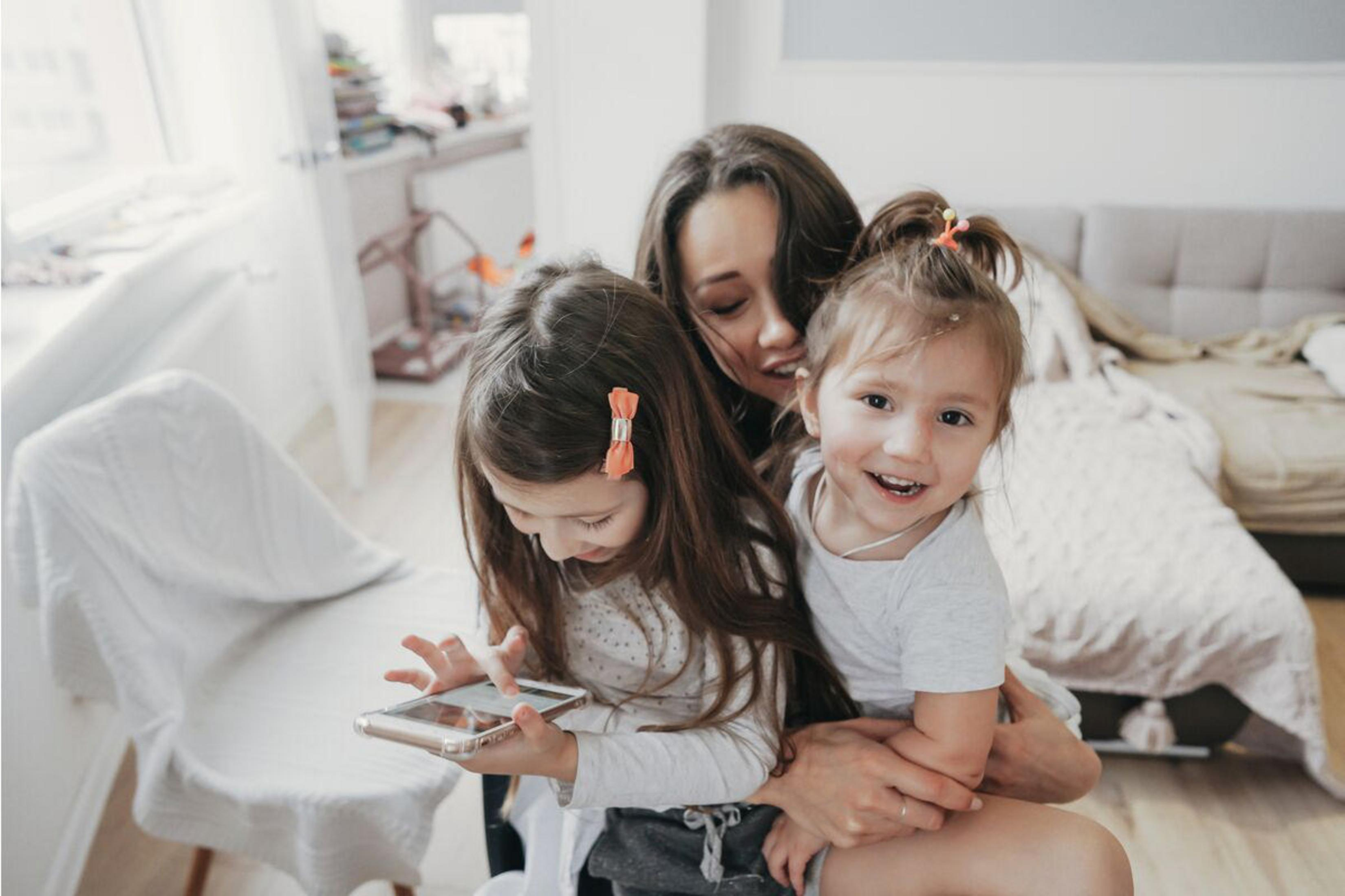 Eine Mutter und zwei Töchter sind in einem Raum. Die Mutter hält die Mädchen. Die Mädchen schauen auf ein Telefon.