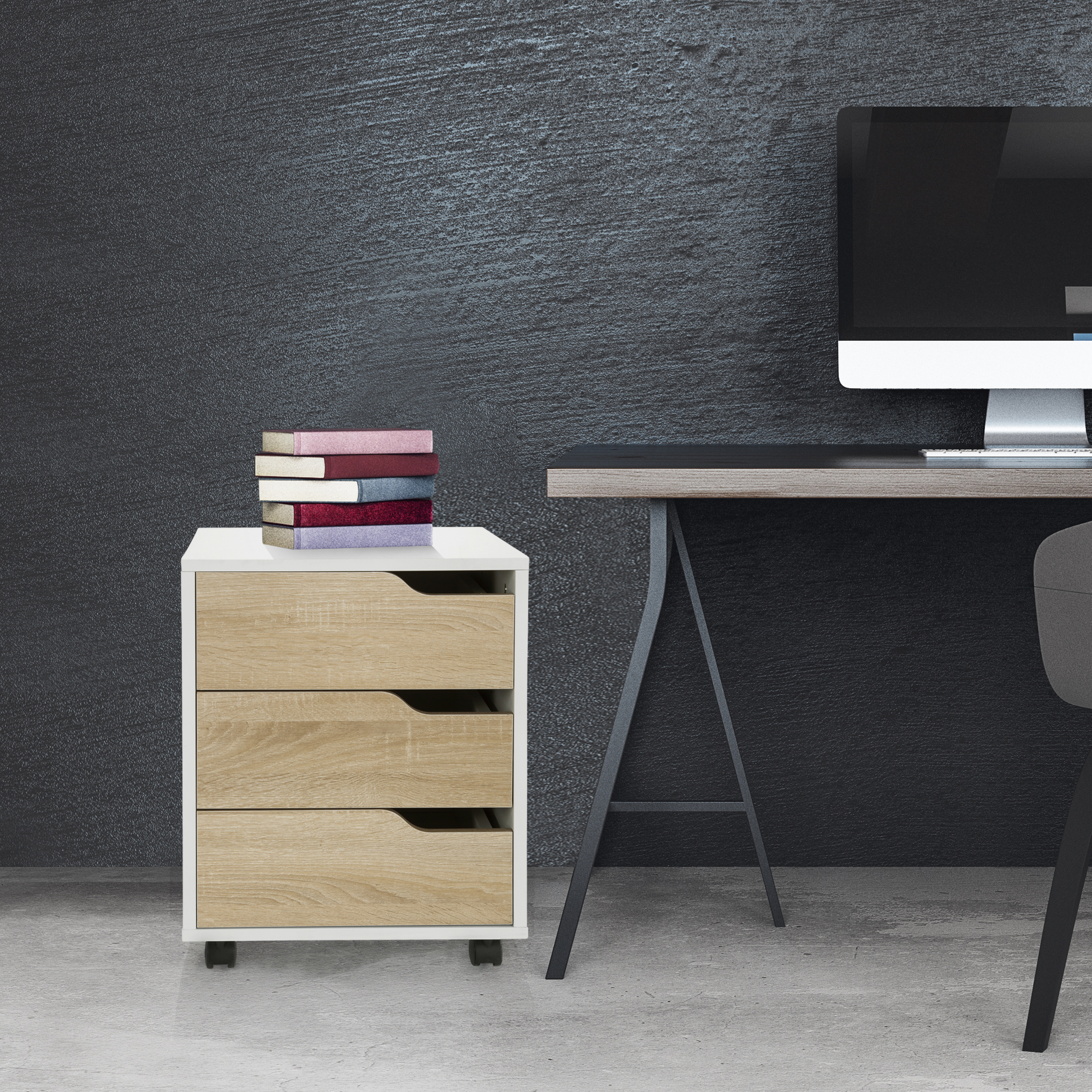 Ein weißer Schrank mit drei Schubladen. Oben sind Bücher, im Hintergrund ein Schreibtisch und Computer.