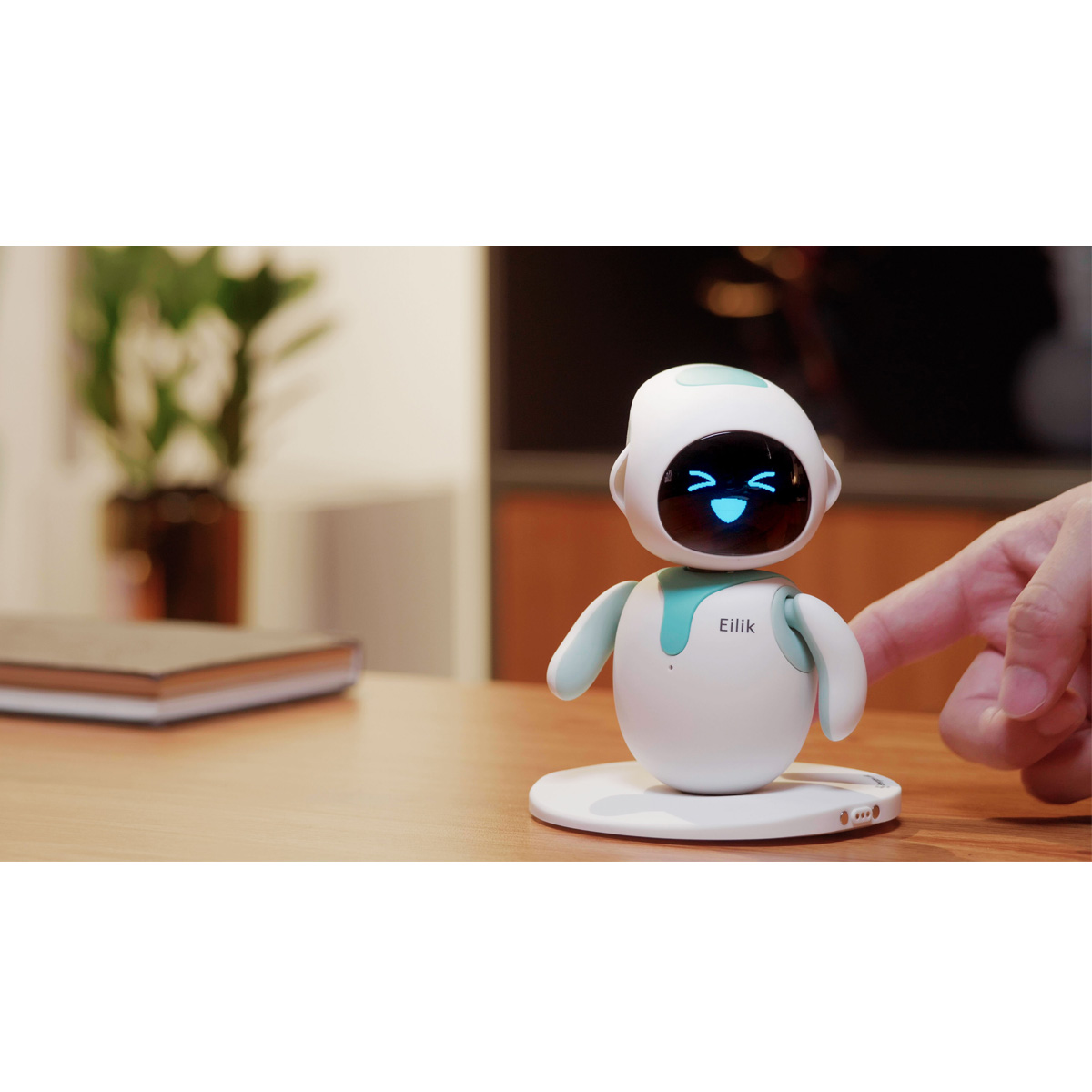 Ein weißer Roboter namens Eilik mit türkisfarbenen Akzenten auf einem Holztisch. Es hat ein blaues, lächelndes Gesicht.