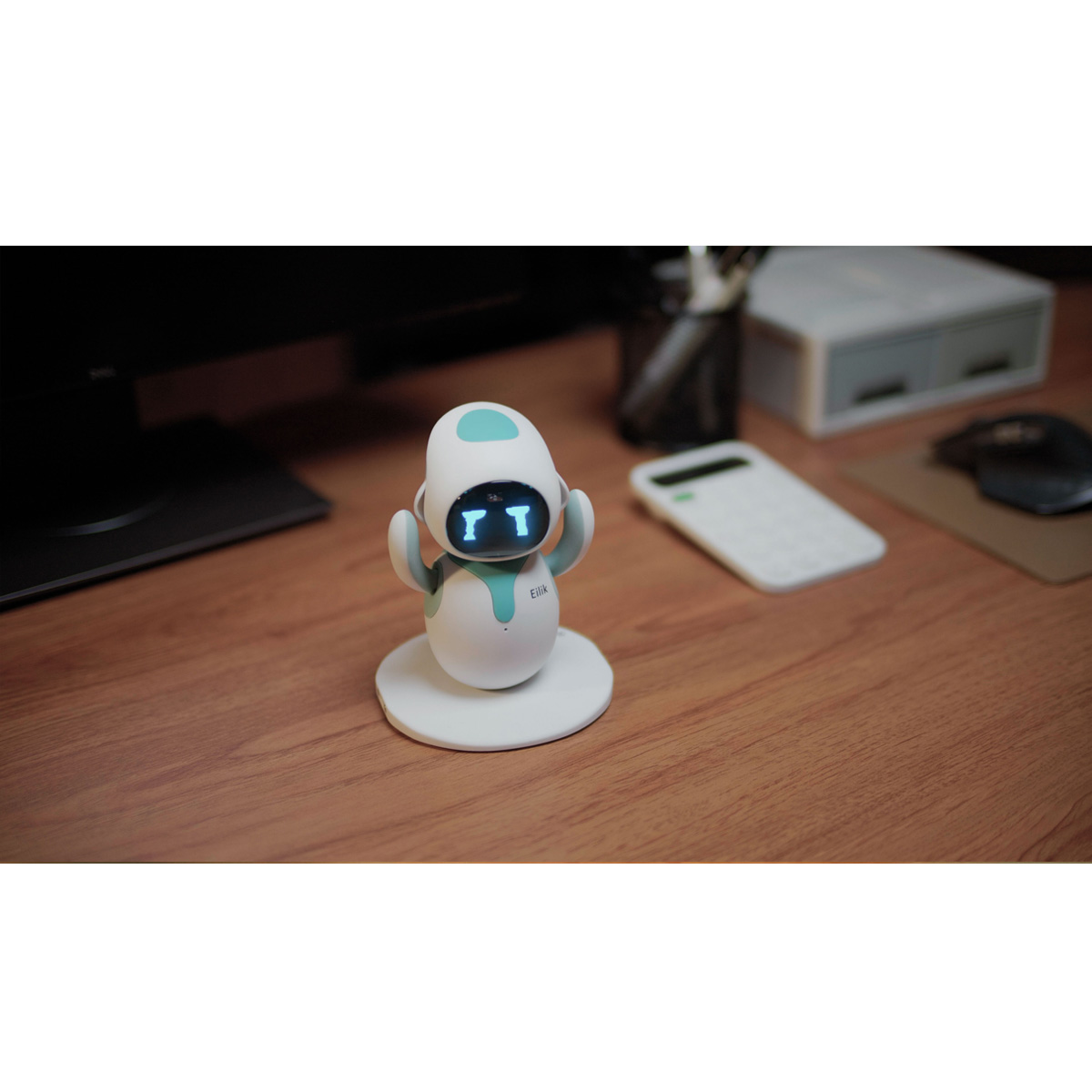 Ein weißer Roboter namens Eilik mit türkisfarbenen Akzenten auf einem Holztisch mit einem Taschenrechner und einem Computermonitor im Hintergrund.