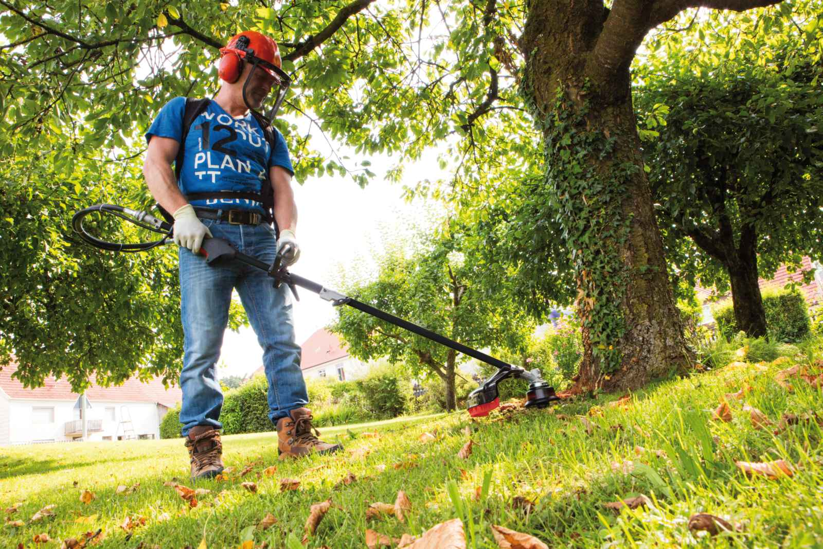 Man gebruikt een grastrimmer in een tuin, draagt veiligheidskleding, bij een boom.
