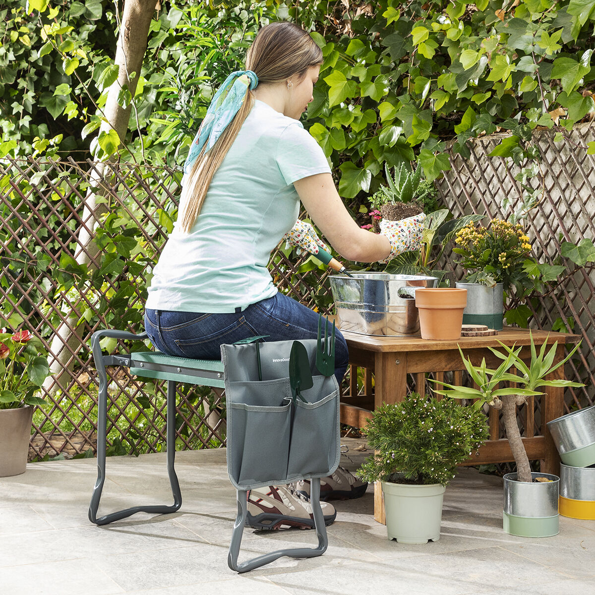 Una donna sta facendo giardinaggio all'aperto, usando una panca con tasche per gli attrezzi. Piante e un tavolo di legno sono sullo sfondo.