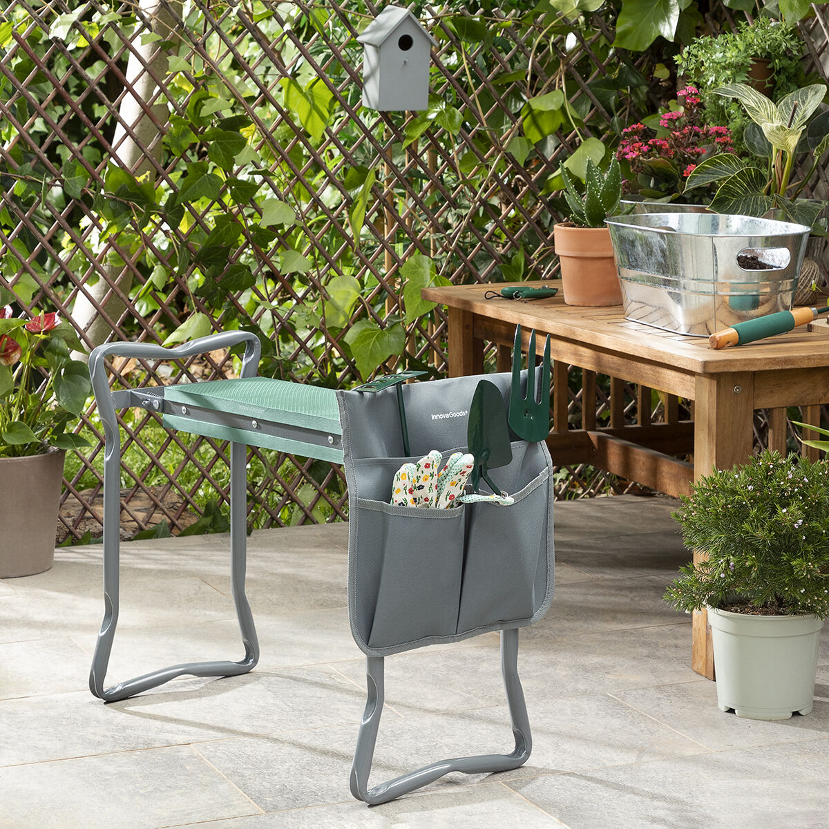 Una panca da giardino verde con attrezzi. Una casetta per uccelli e piante sullo sfondo. Ambiente esterno soleggiato.