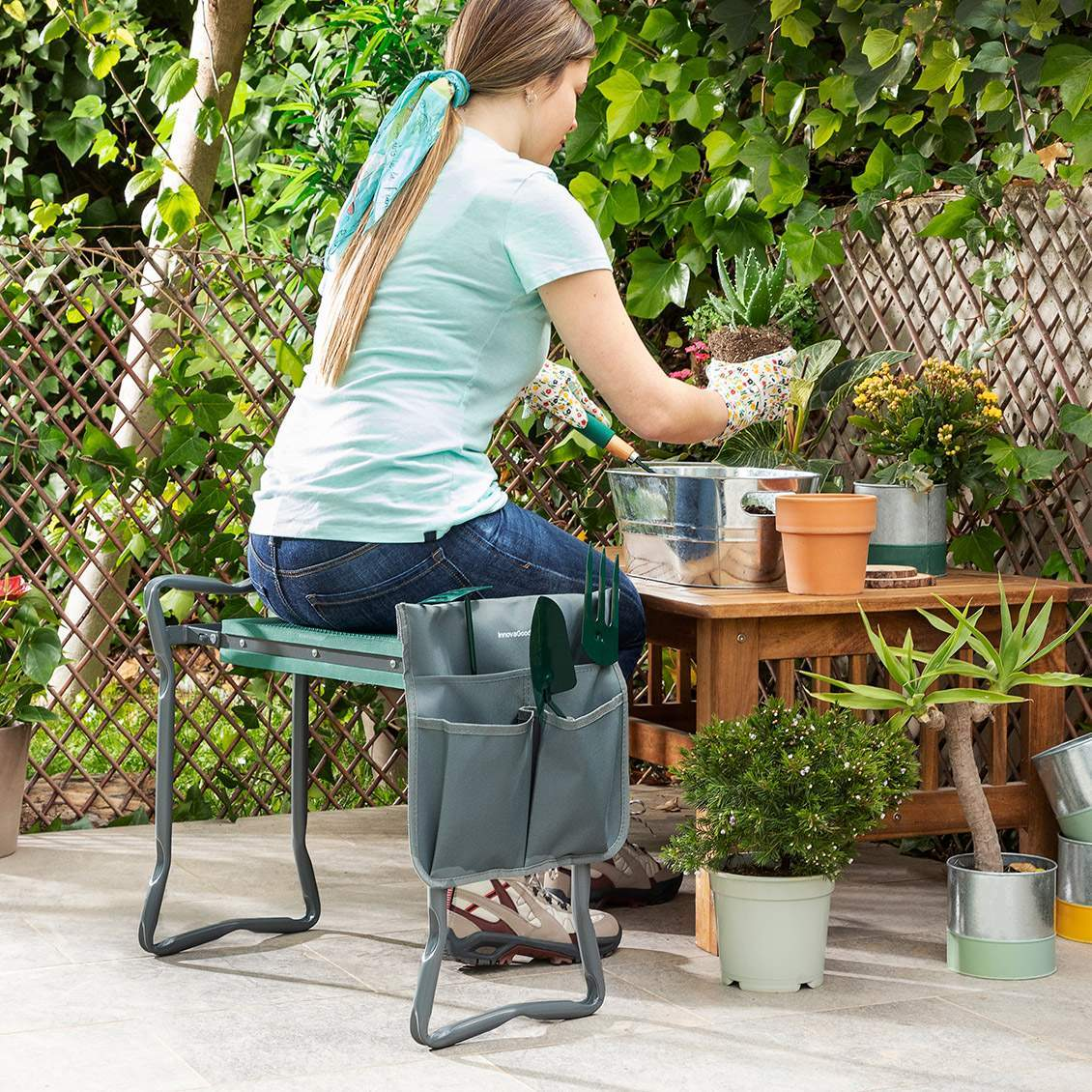 Donna che fa giardinaggio, seduta su uno sgabello verde con attrezzi. Piante, tavolo in legno e recinzione sullo sfondo.