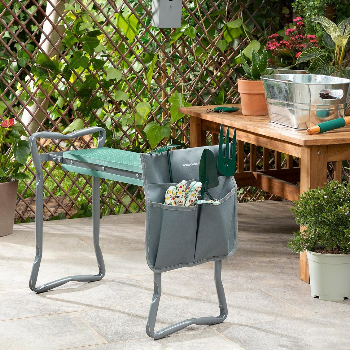 Una donna in giardino è inginocchiata su un sedile verde con attrezzi da giardinaggio nelle tasche e un tavolo di legno sullo sfondo.