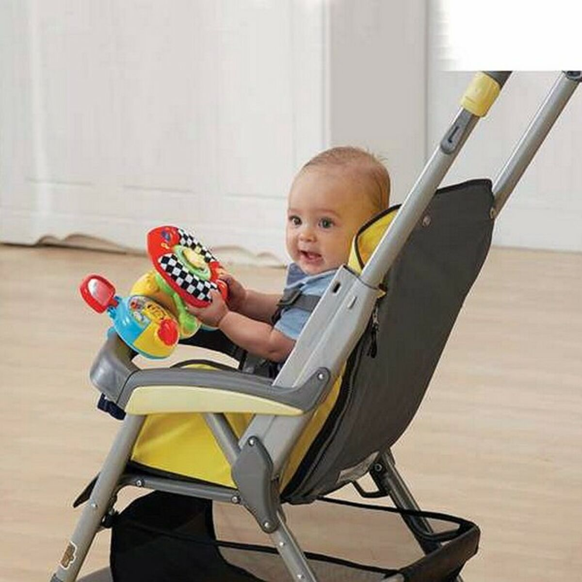 Un bebé en un cochecito jugando con un volante de juguete. El cochecito es gris y amarillo con un bebé sonriente.