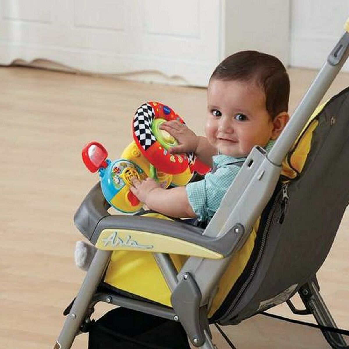 Un bebé en un cochecito con un volante de juguete. El bebé sonríe. El cochecito es amarillo y gris.