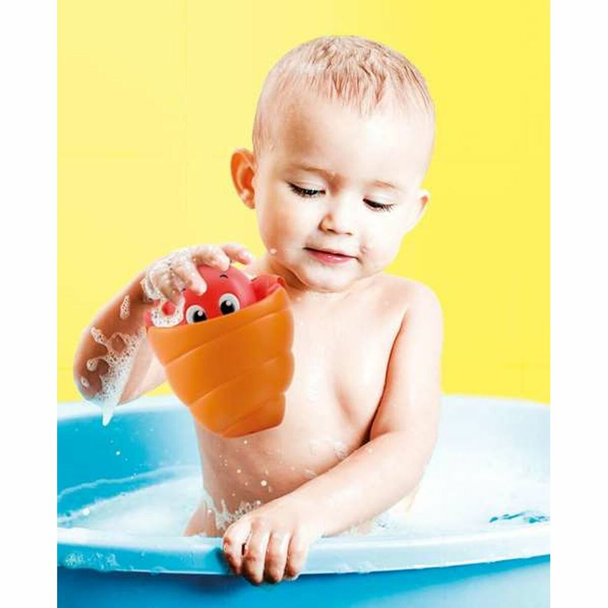 Un bebé está en una bañera jugando con un juguete rojo y naranja frente a un fondo amarillo.