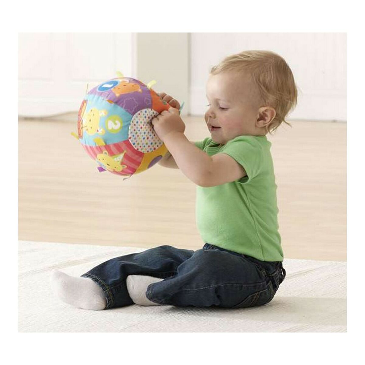 Un bebé se sienta y sostiene una pelota colorida con formas y texturas. El bebé viste una camisa verde y jeans.