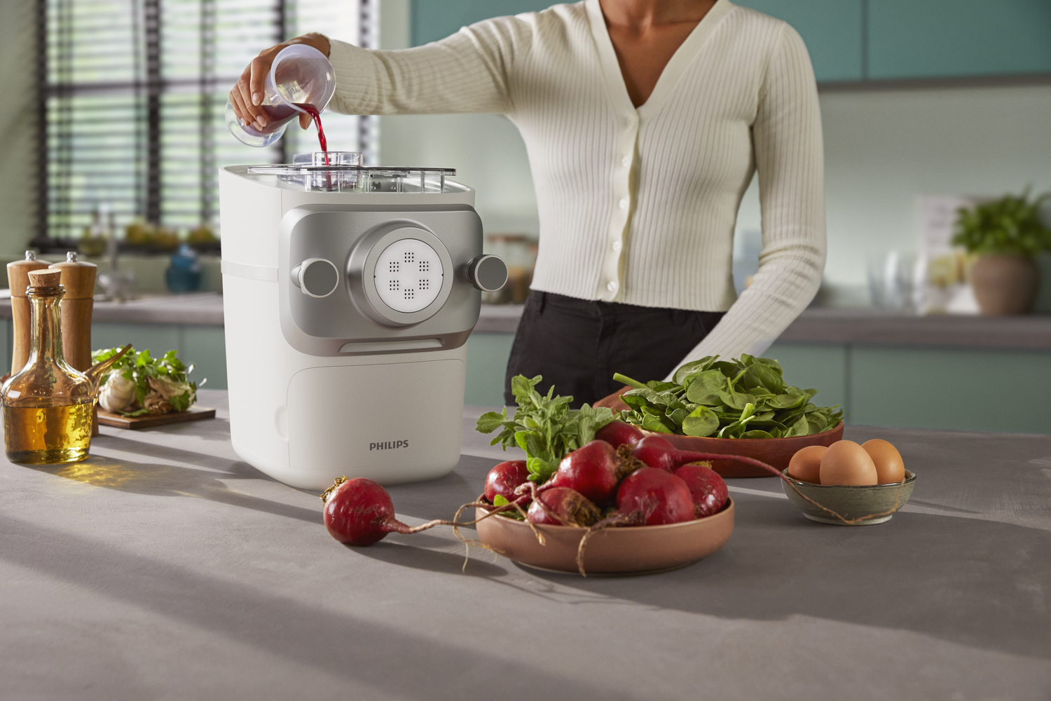 Osoba wlewa płyn do białej maszynki do makaronu. Na blacie znajdują się warzywa i inne składniki.