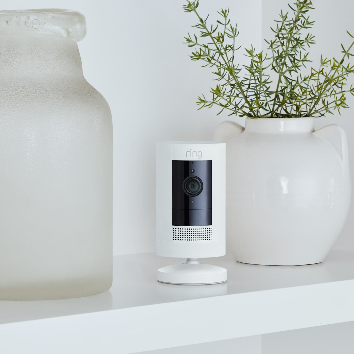 Biała kamera bezpieczeństwa na białej półce. Obok szklany słoik i wazon z rośliną.