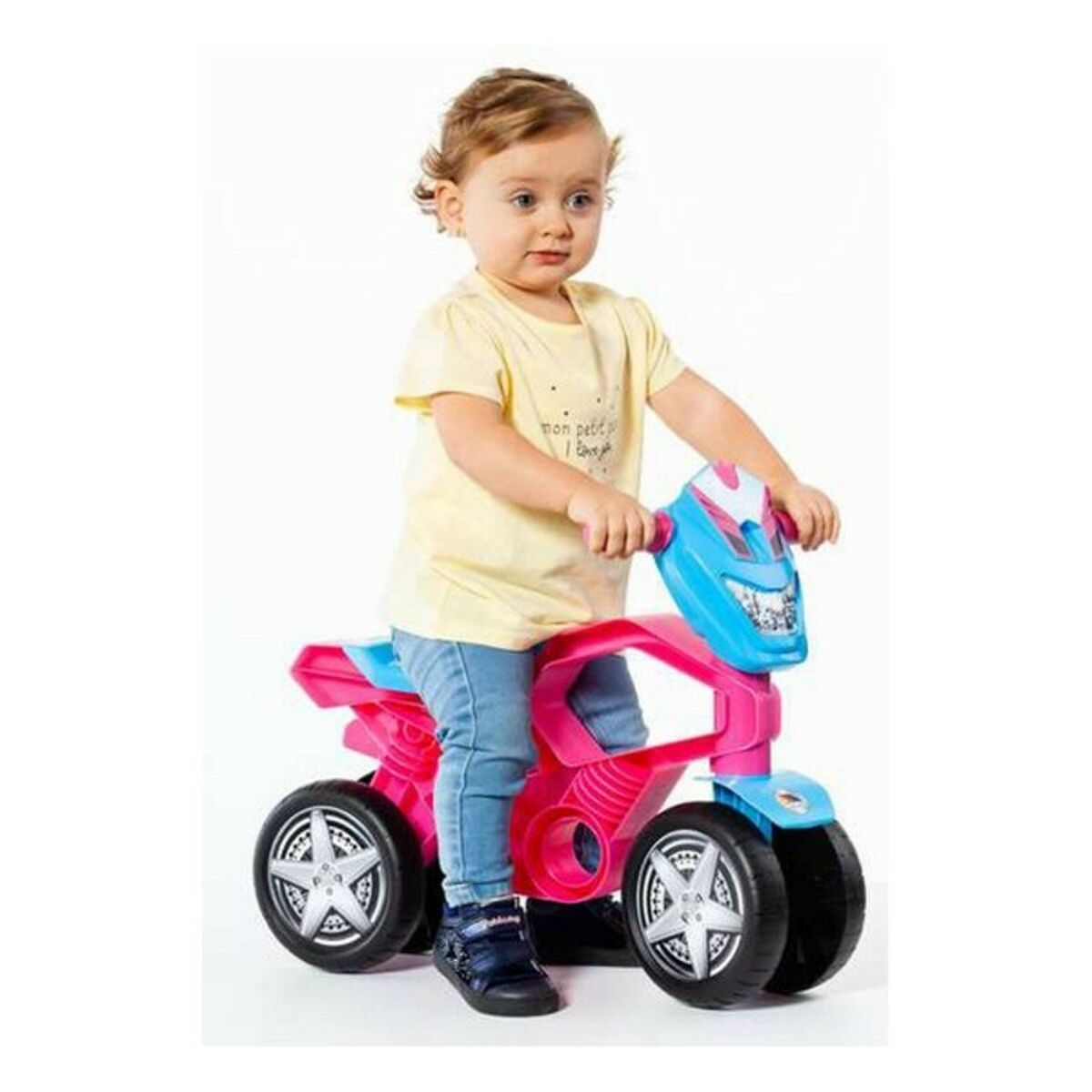 Un bambino piccolo è seduto su una moto giocattolo rosa, blu e nera, sfondo bianco.