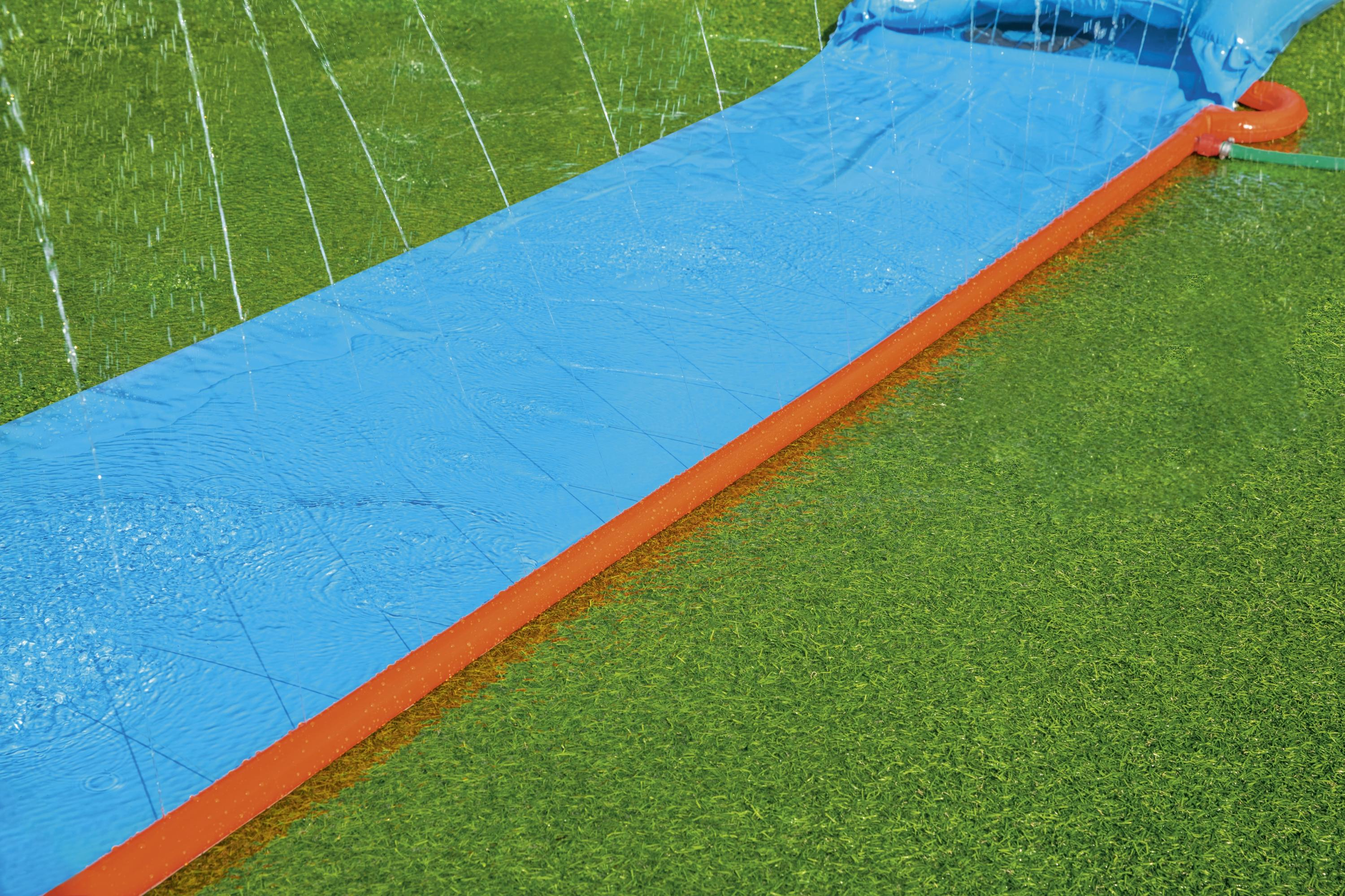 Zjeżdżalnia wodna na zielonej trawie. Niebieska zjeżdżalnia i pomarańczowa krawędź. Biała woda płynie.