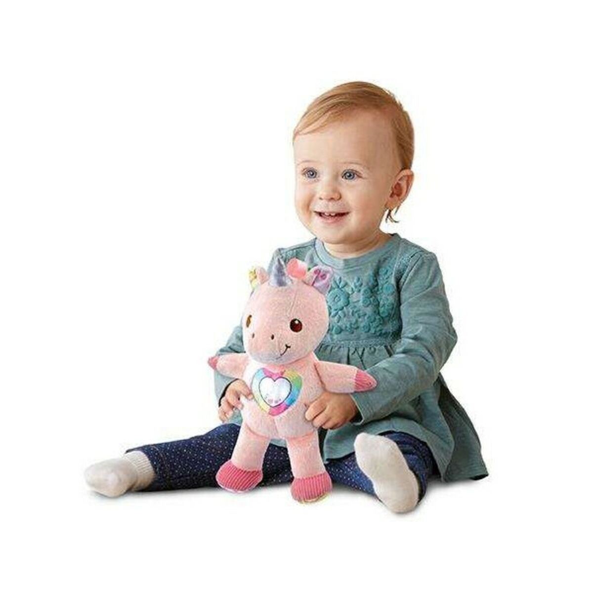 Bebé niña sosteniendo un peluche de unicornio rosa. Fondo blanco, sonriendo, con vestido azul.