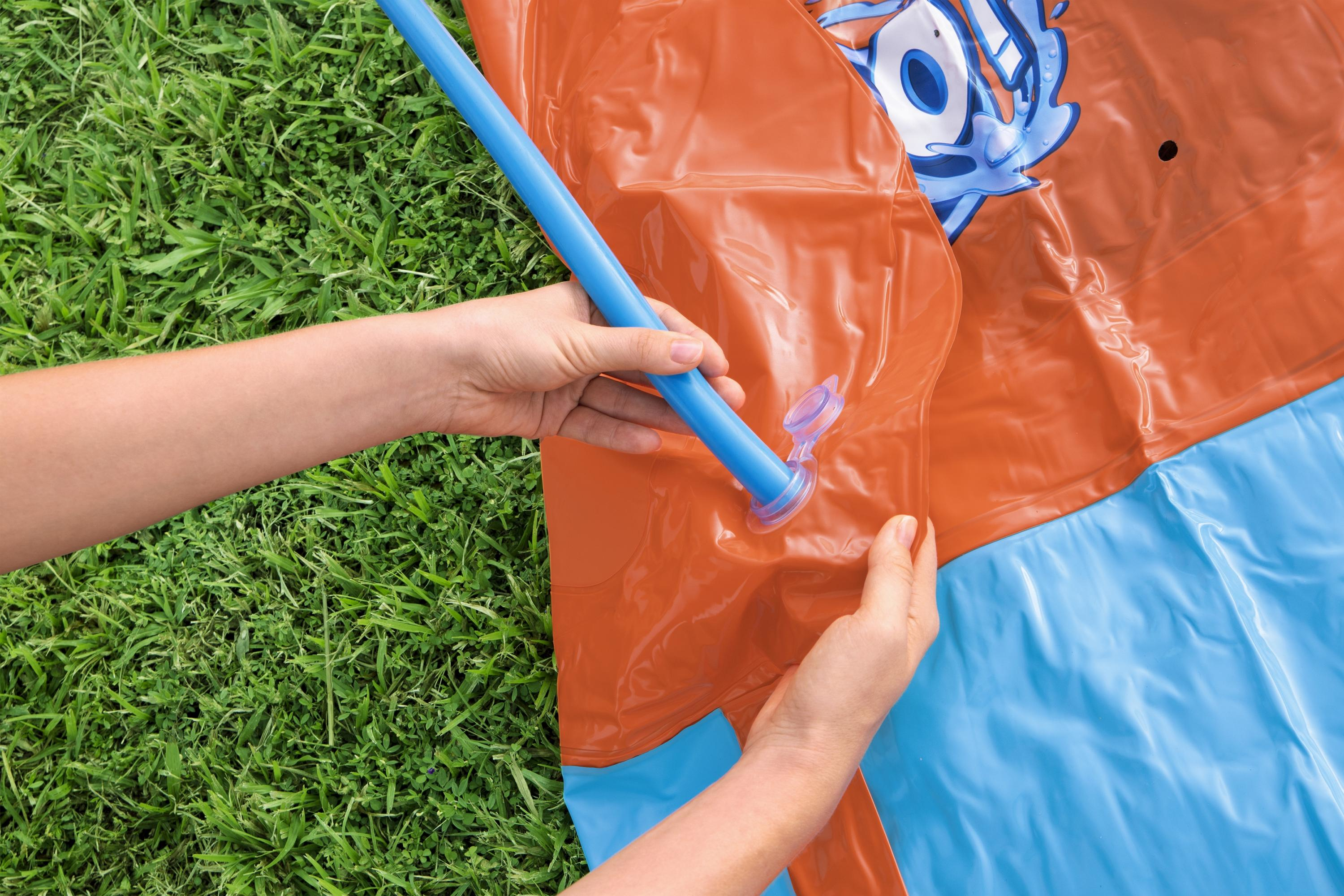 Ręka mocuje niebieską rurę do pomarańczowej zjeżdżalni wodnej. Zielona trawa w tle.