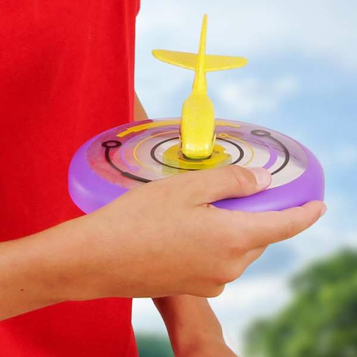 Una mano sostiene un avión de juguete amarillo y morado. Una camisa roja en el fondo.