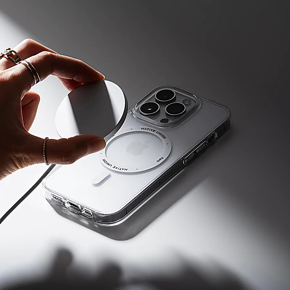 Mano che tiene un pad di ricarica bianco, sopra un telefono in una custodia trasparente. Il telefono è su una superficie chiara.
