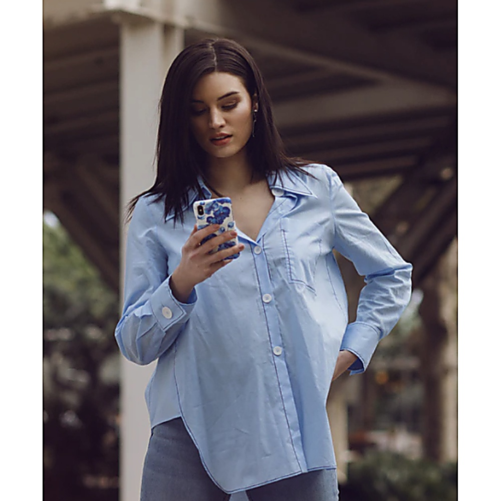 Donna in camicia blu che guarda il telefono, mani sui fianchi, sfondo esterno.