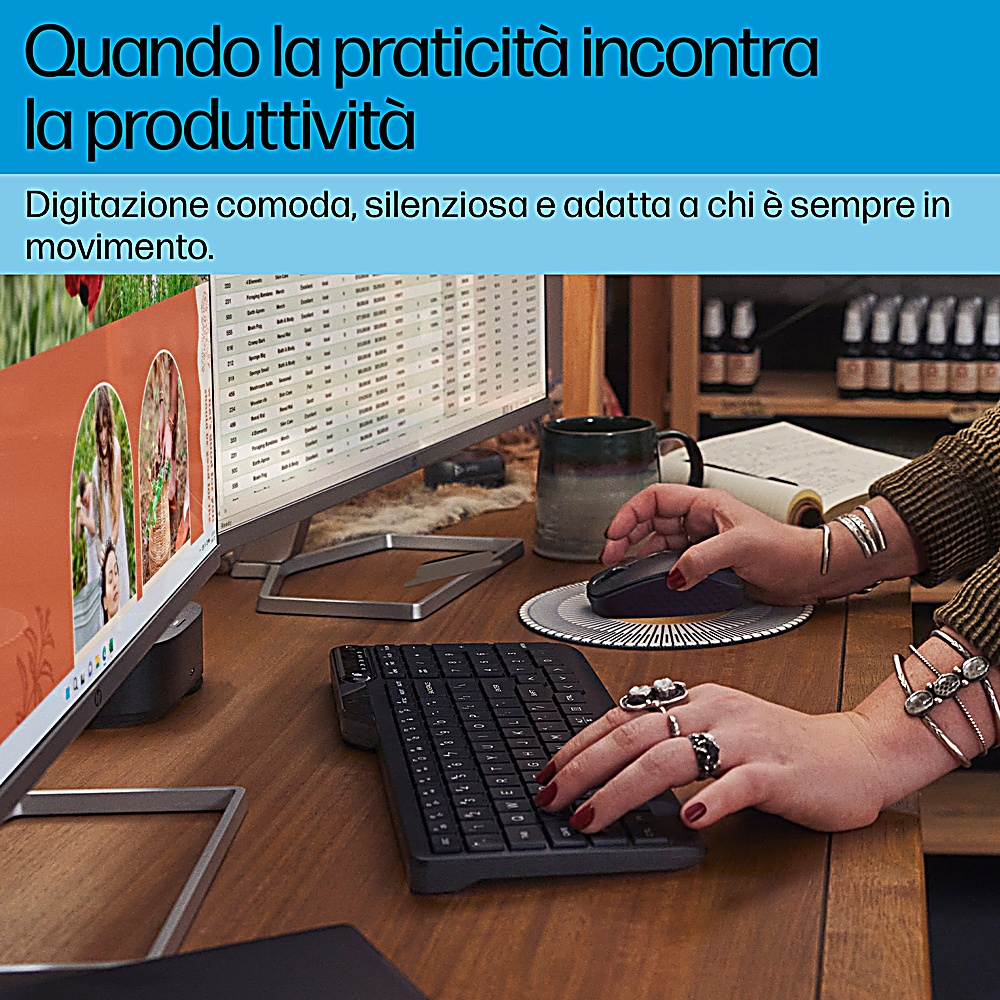 Mani che usano una tastiera e un mouse wireless neri su una scrivania di legno. Un monitor è sullo sfondo.