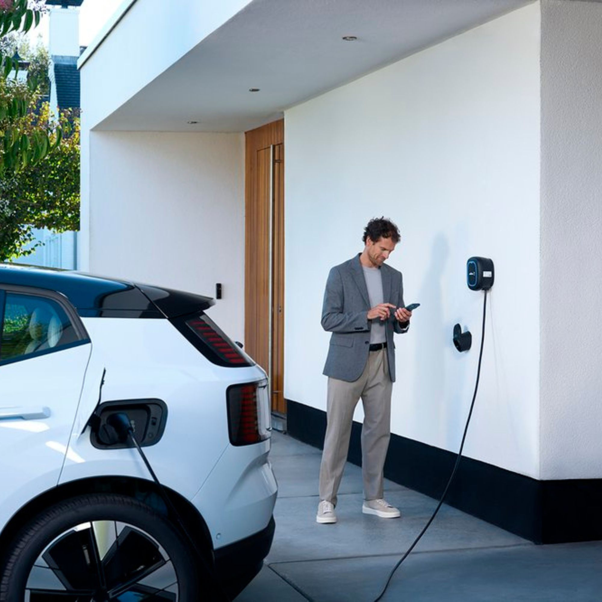 Ein Mann steht in der Nähe eines weißen Autos und eines Wandladegeräts. Das Auto ist eingesteckt. Der Mann hält ein Telefon.