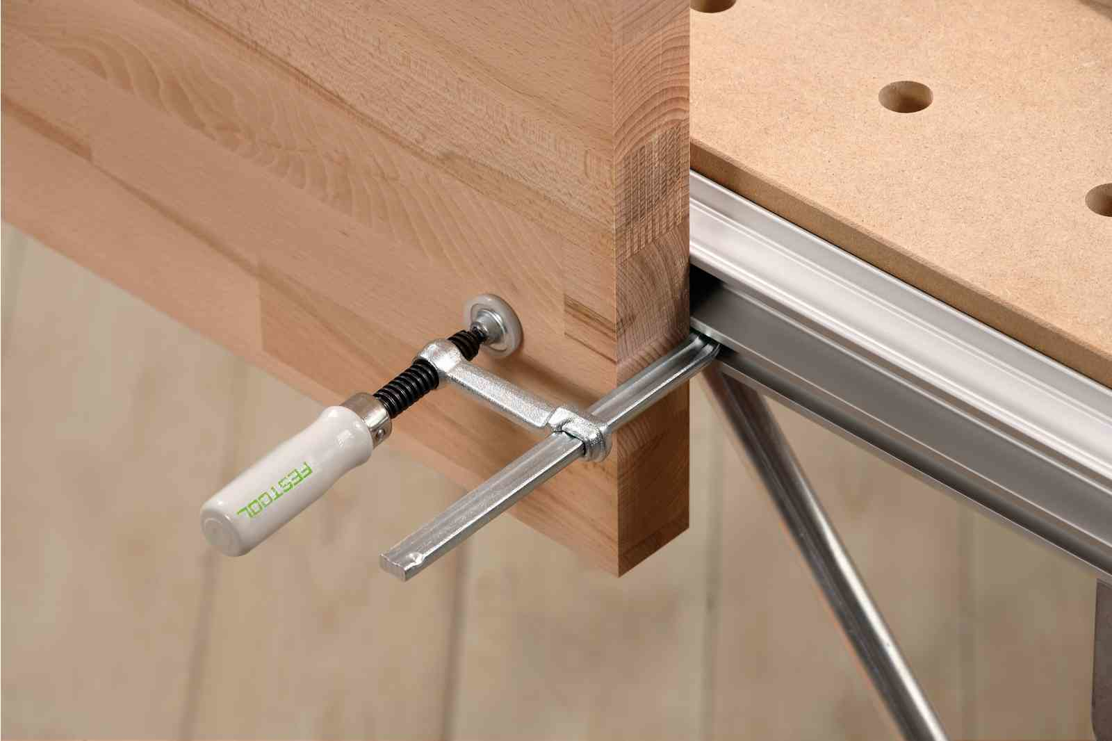 Zacisk Festool mocujący drewniany element do stołu warsztatowego. Zacisk jest srebrny i biały, a stół drewniany.