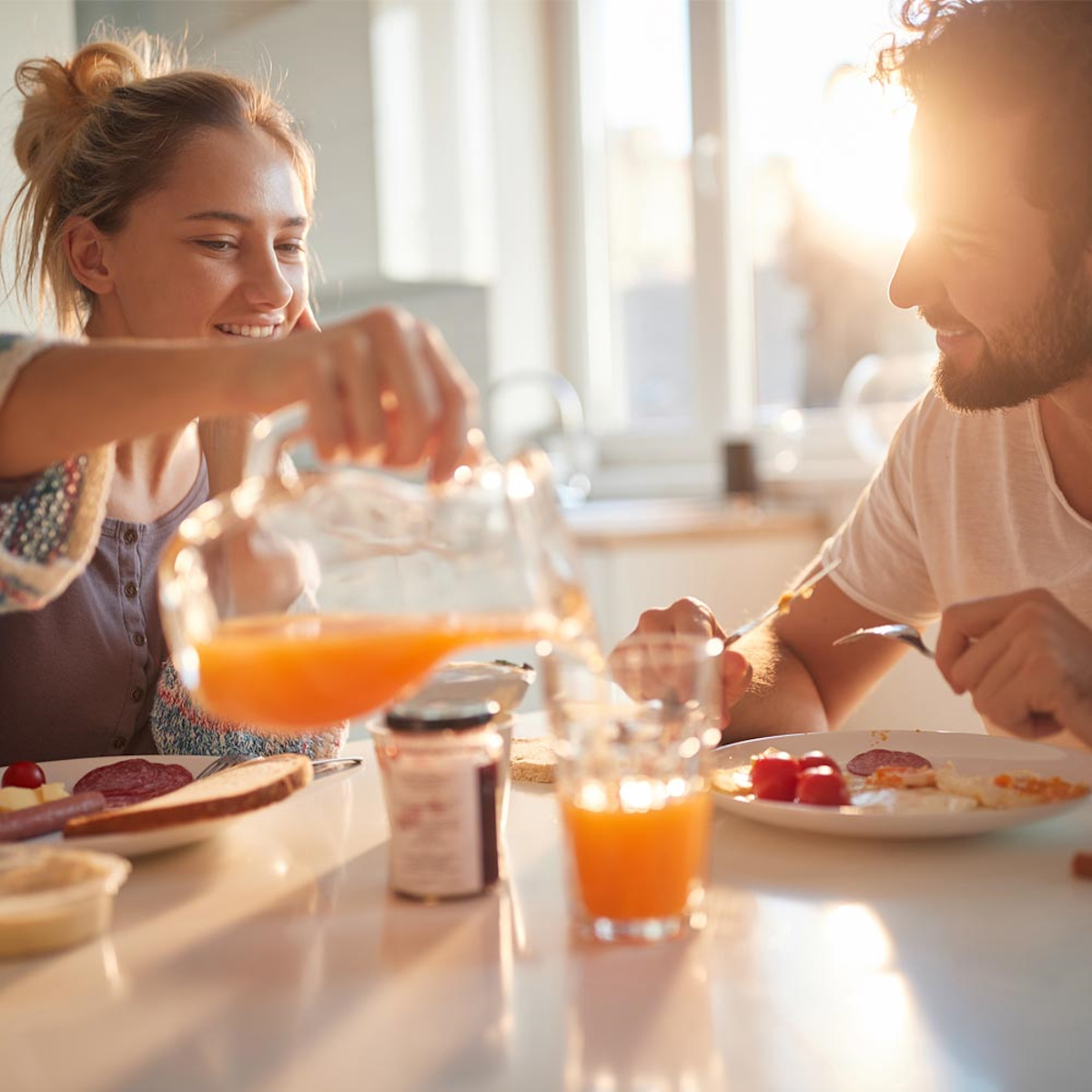 Paar beim Frühstück. Frau gießt Saft ein. Essen auf dem Tisch: Brot, Wurst, Marmelade. Sonniger Küchenhintergrund.