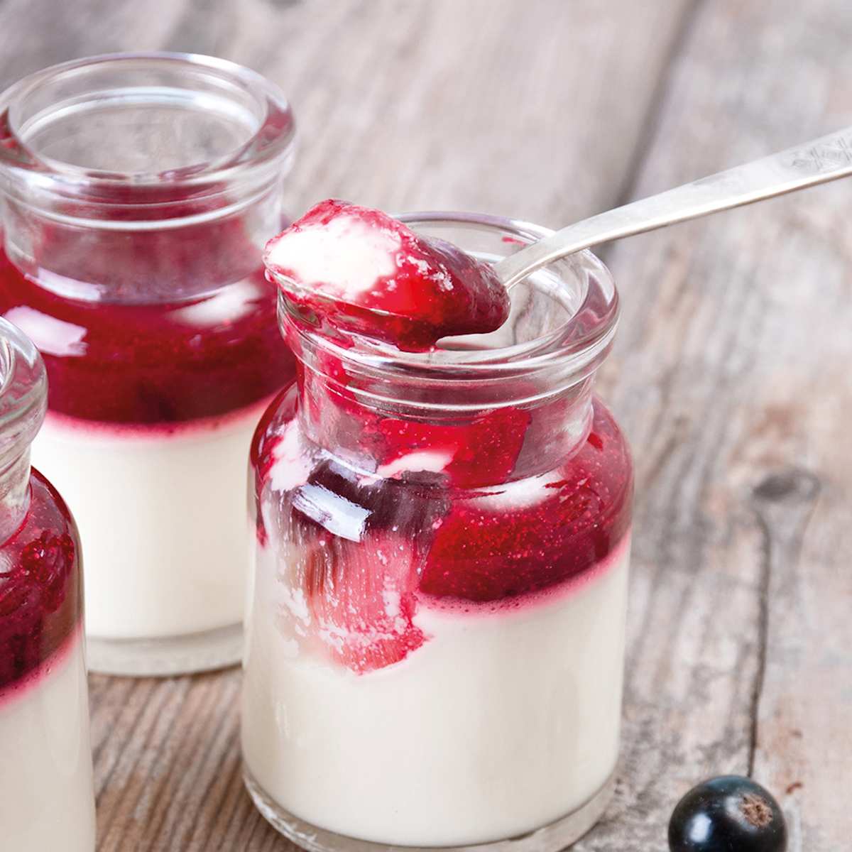 Gläser mit Schichten aus weißer Creme und roter Beerensauce. Löffel in einem Glas. Holzoberfläche, schwarze Beeren.