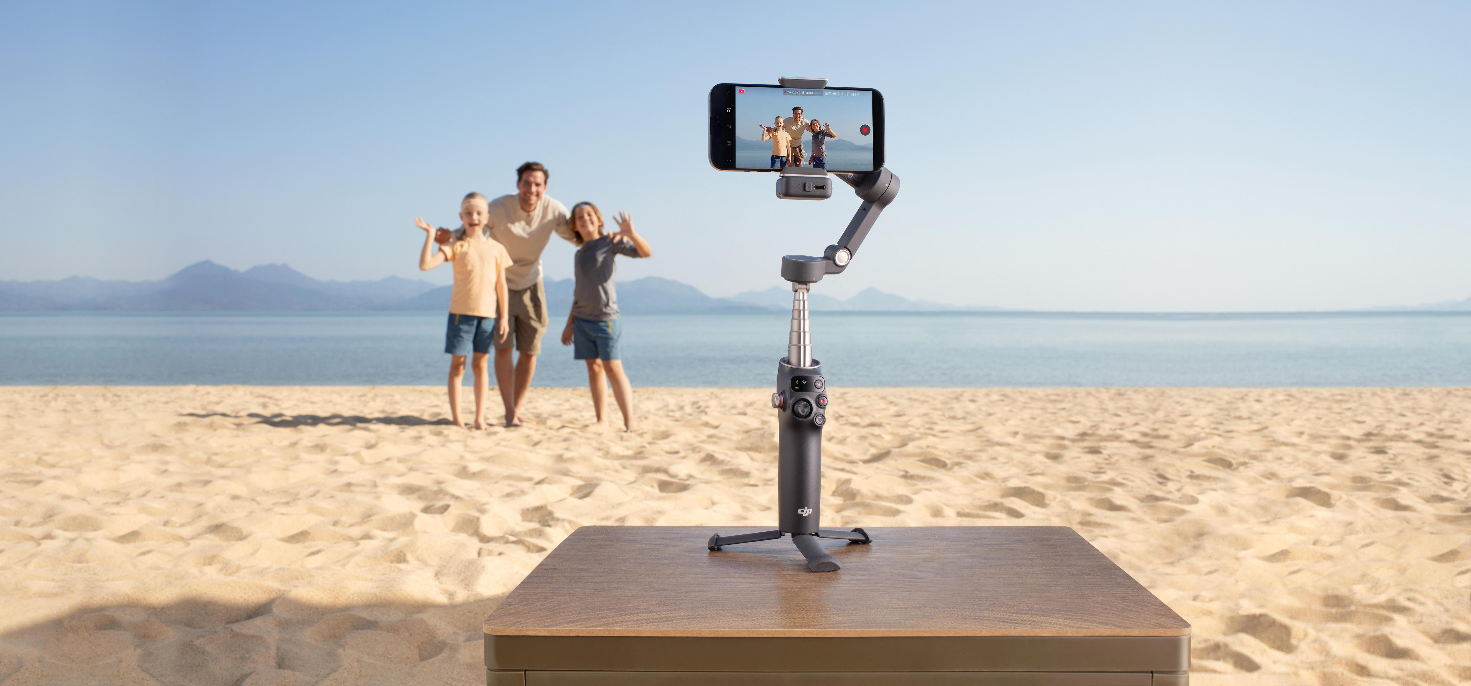 DJI Osmo Mobile na stole, nagrywający rodzinę na plaży. Błękitne niebo, morze w tle.