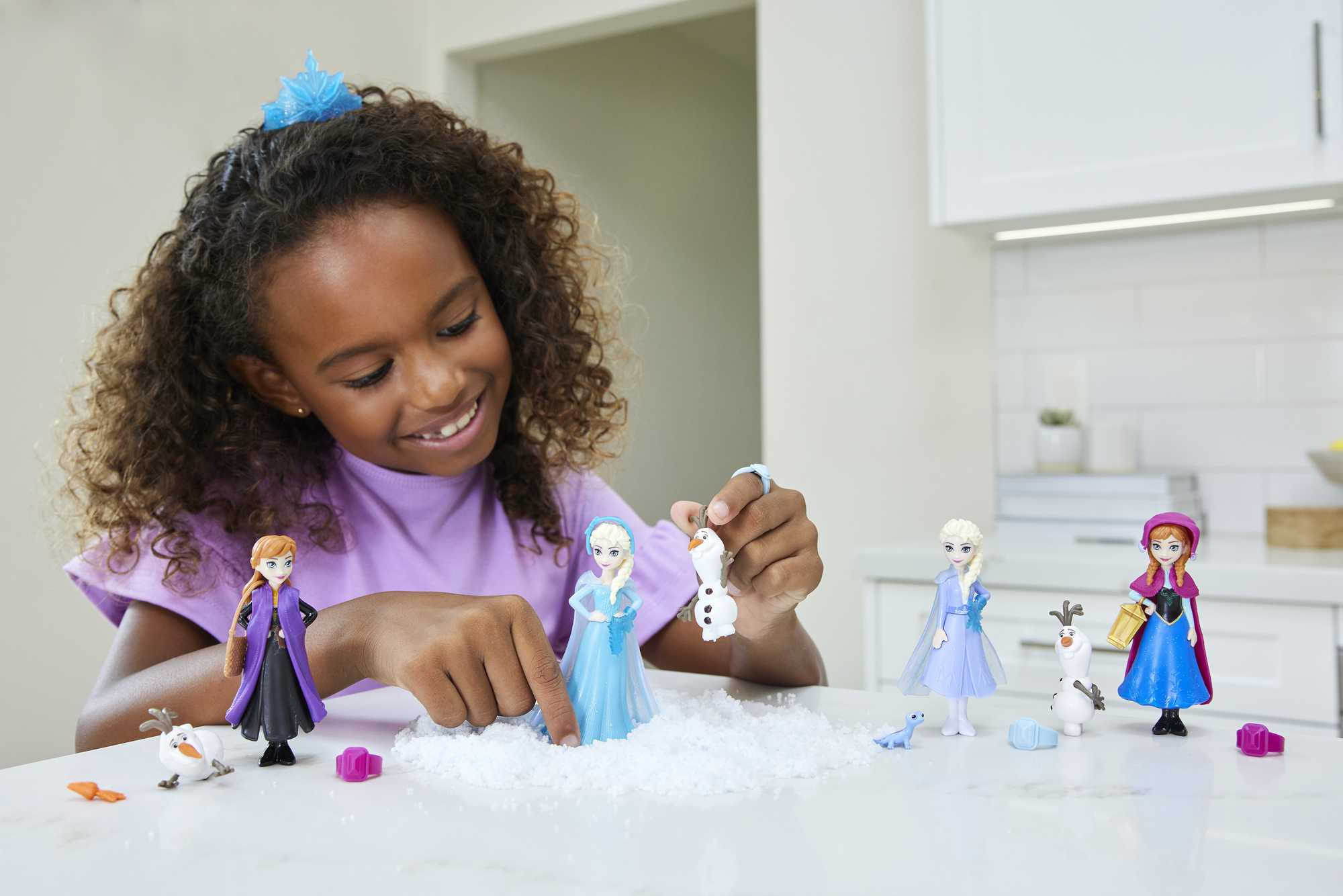 Una bambina gioca con le figure di Frozen. La scena è su un tavolo bianco con neve, con le figure di Anna ed Elsa.