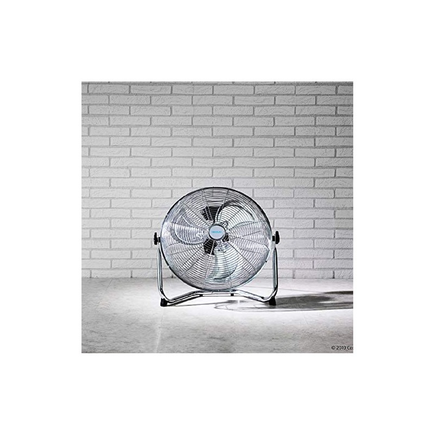 Srebrny wentylator elektryczny na szarej podłodze przed białą ceglaną ścianą. Okrągły, metalowa rama.