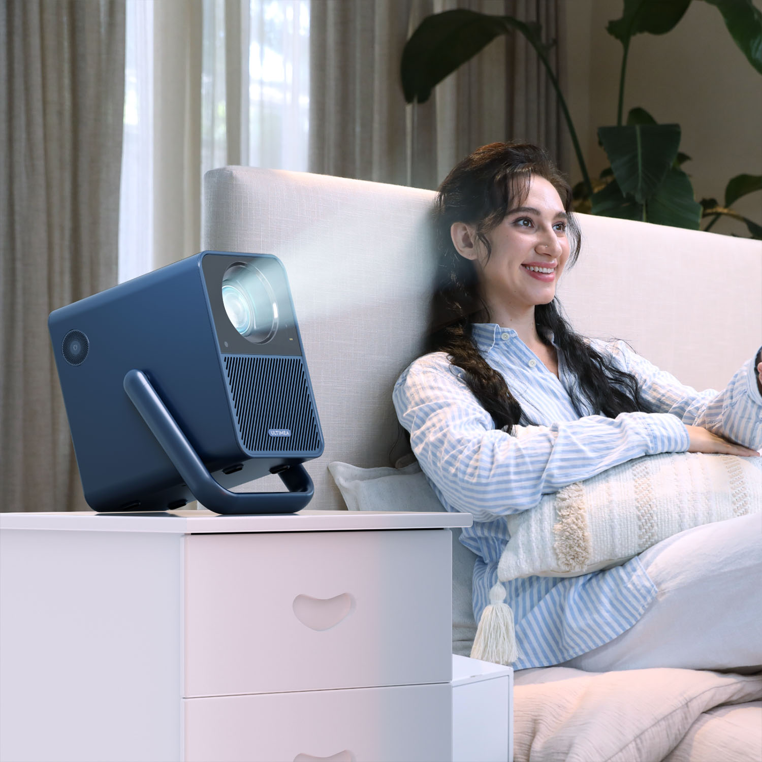 Une femme sourit assise sur un lit. Un projecteur bleu est sur une table de chevet, projetant une image.