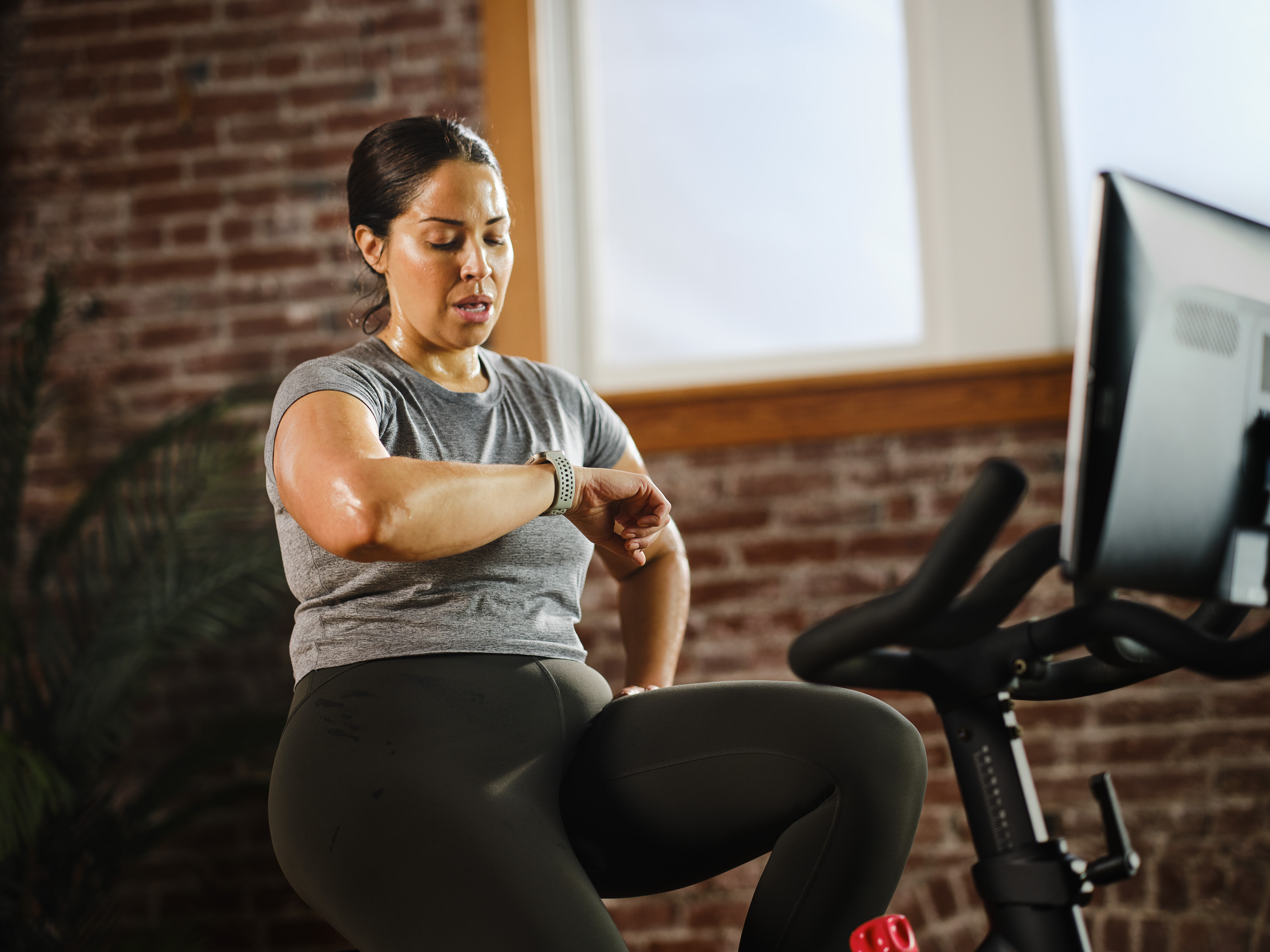 Kobieta ćwiczy na rowerze, patrząc na swój smartwatch. Pokój z ceglaną ścianą.
