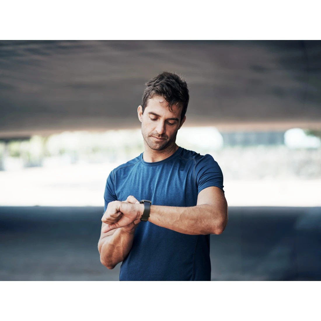 Uomo che guarda uno smartwatch. Indossa una camicia blu. L'ambientazione è sotto un ponte.