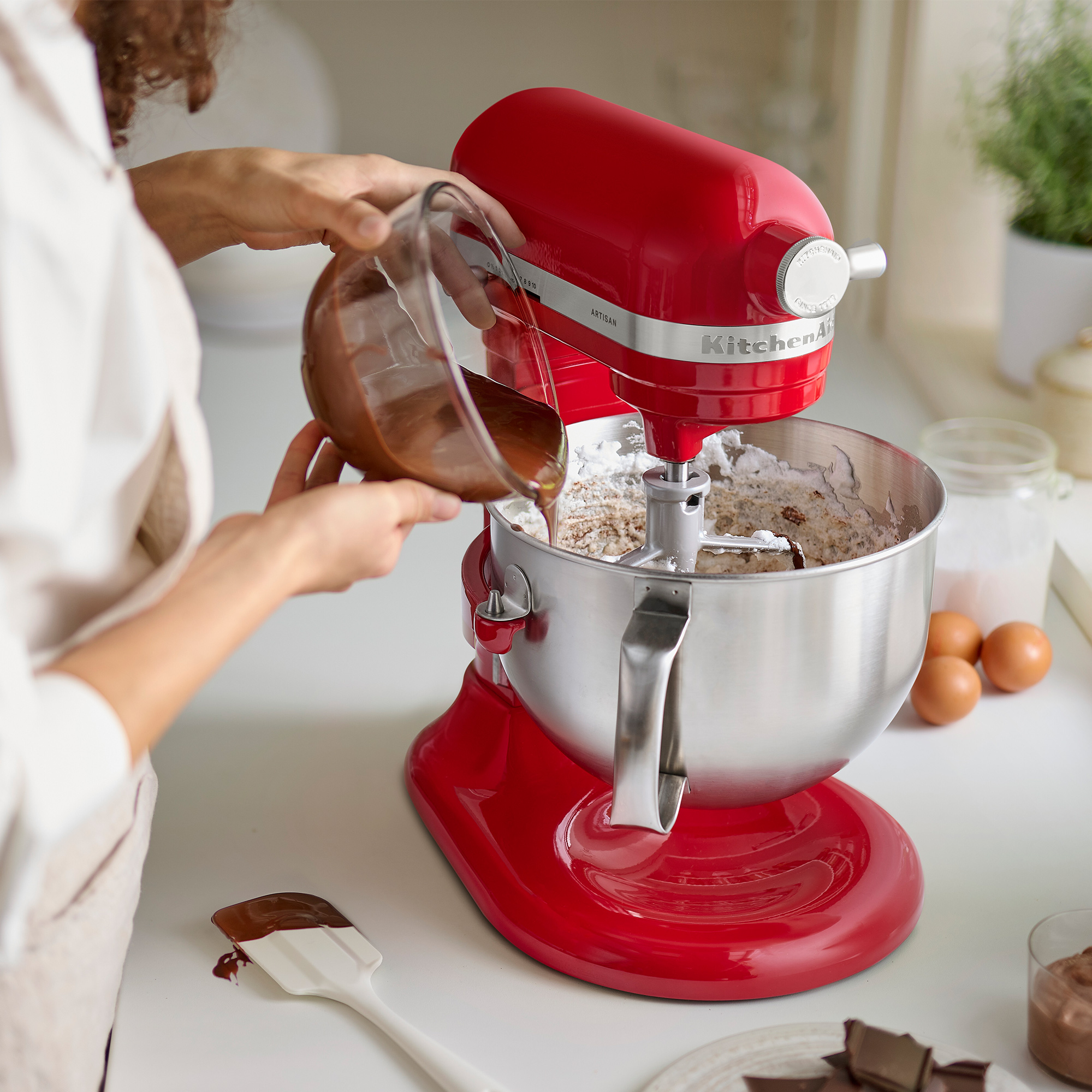 Una persona versa l'impasto al cioccolato in un mixer KitchenAid rosso. Altri ingredienti e una spatola sono vicini.