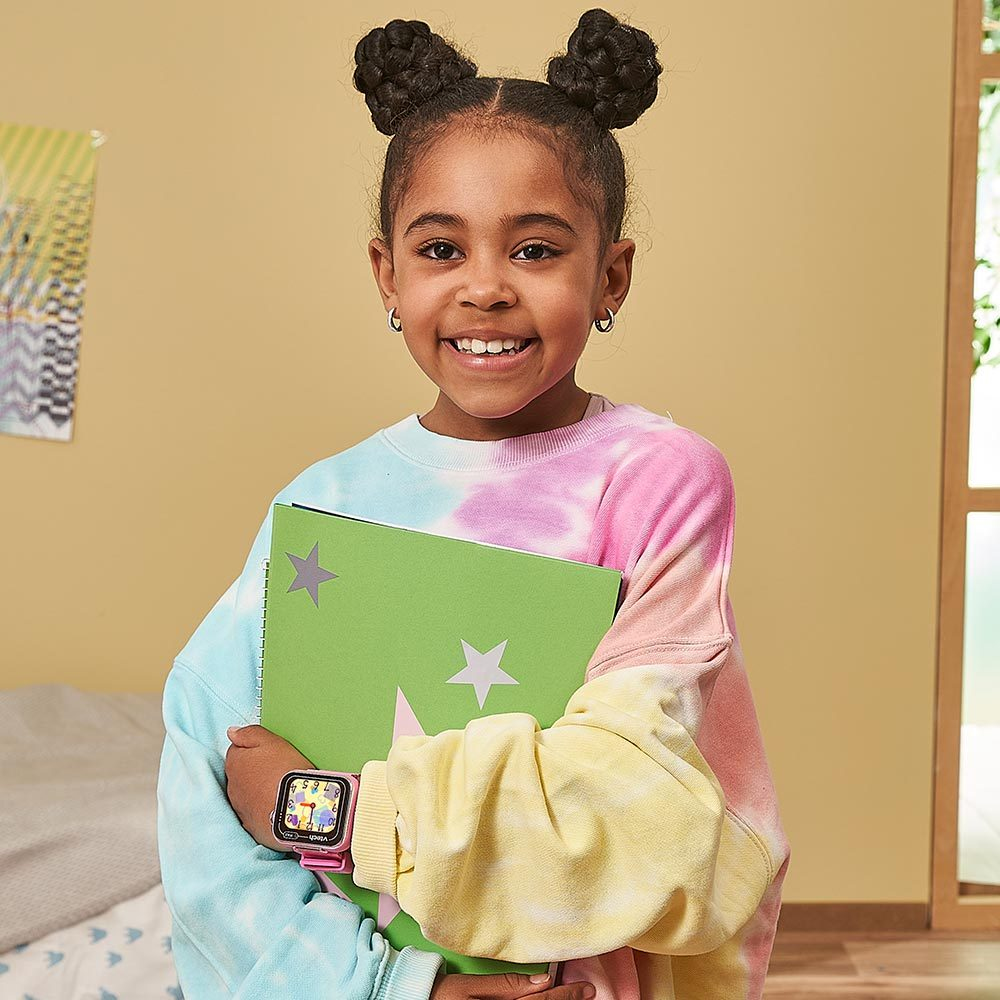 Niña con moños sosteniendo un cuaderno verde. Lleva un suéter teñido y un reloj inteligente rosa, sonriendo.