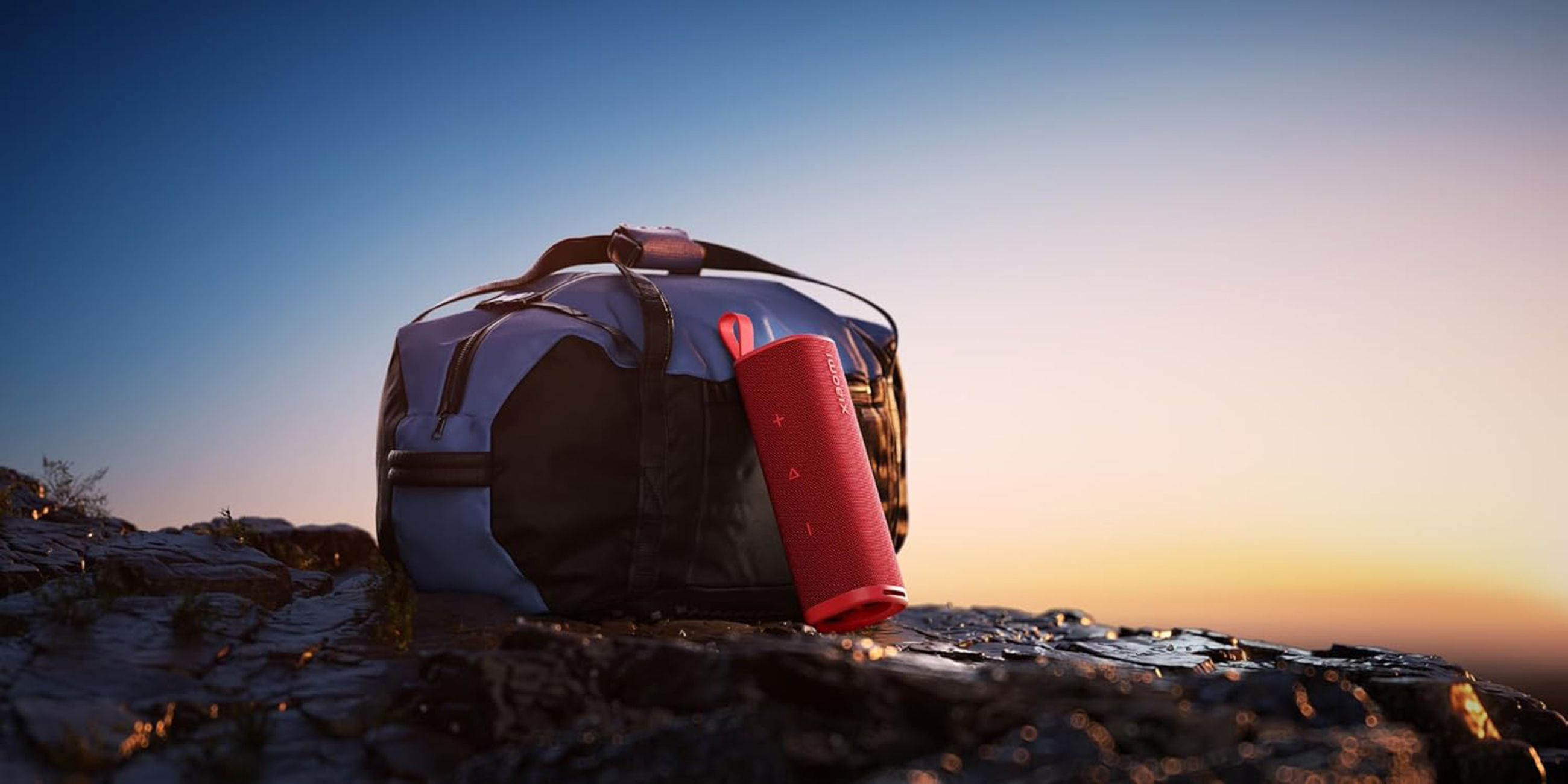 Czerwony głośnik i niebieska torba na skale, z zachodem słońca w tle.