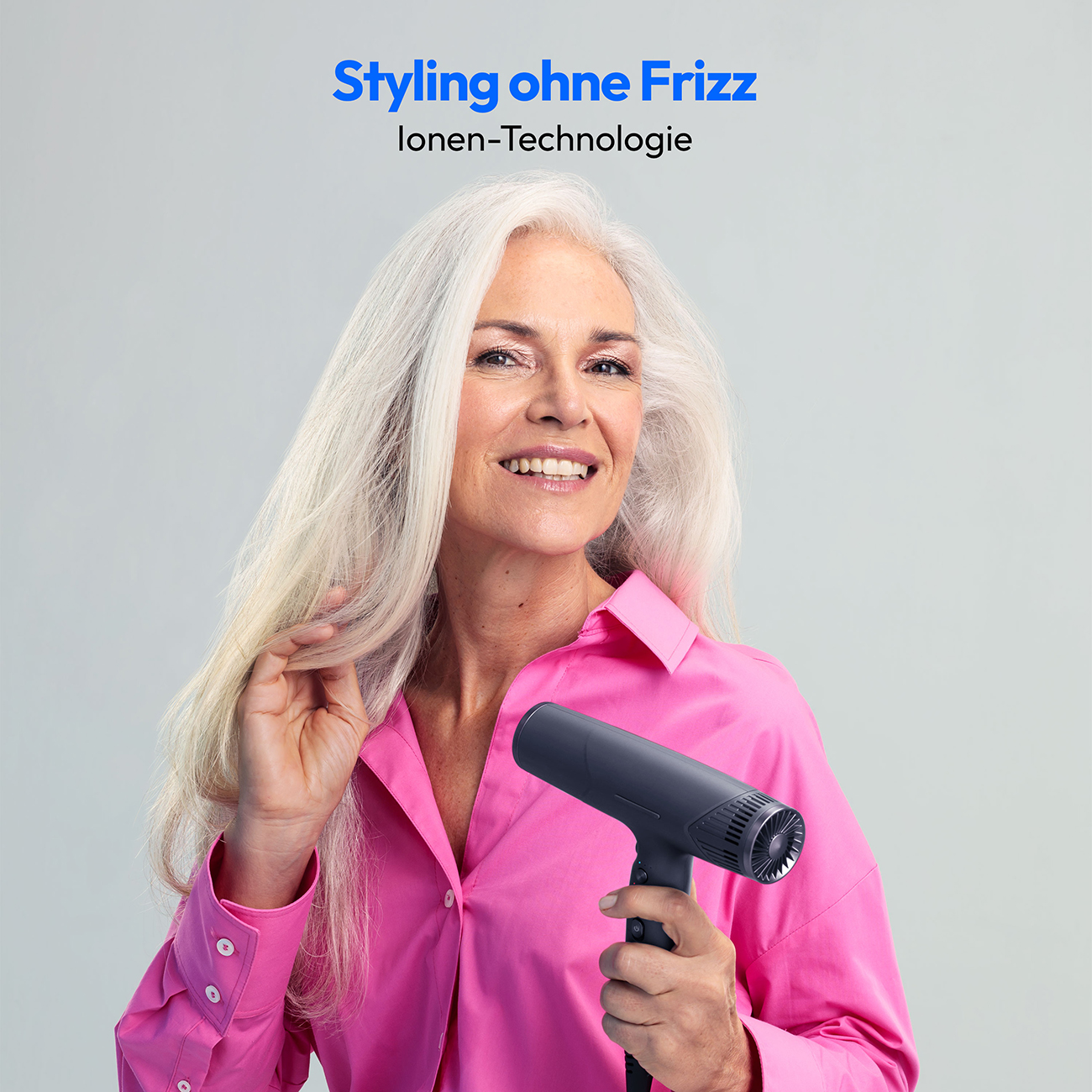 Kobieta trzymająca czarną suszarkę do włosów, uśmiechnięta, różowa koszula, jasne tło.
