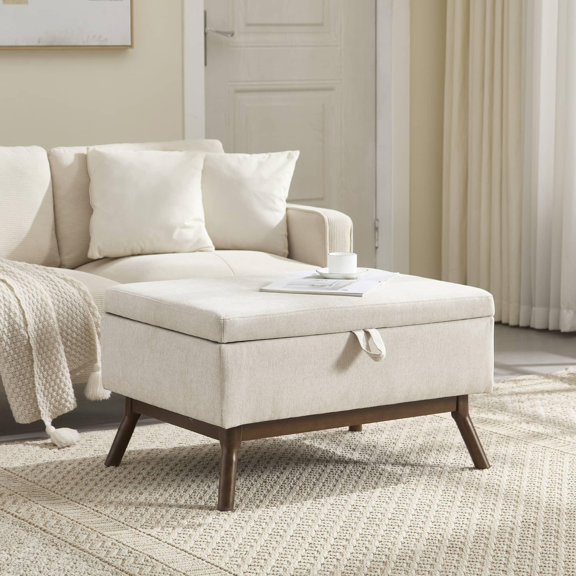 Eine beige Ottomane in einem Wohnzimmer mit Sofa, Teppich und einem Beistelltisch mit Tasse und Buch.