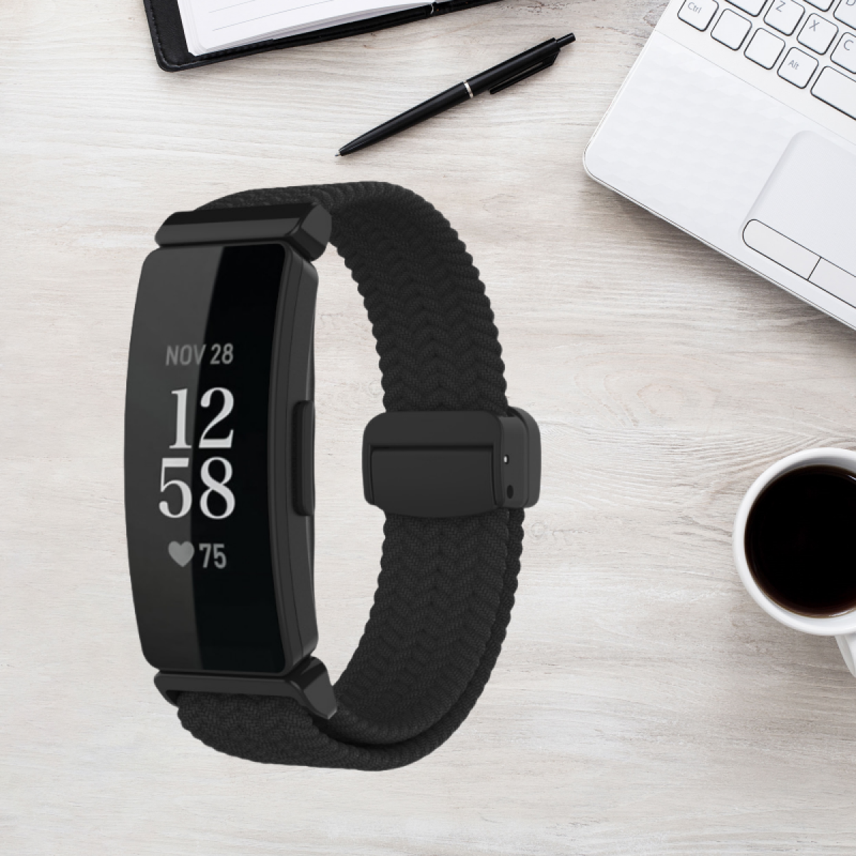 Fitnesstracker auf einem Holztisch. Anzeige zeigt Zeit, Datum und Herzfrequenz. Auch Stift, Notizbuch und Tasse.