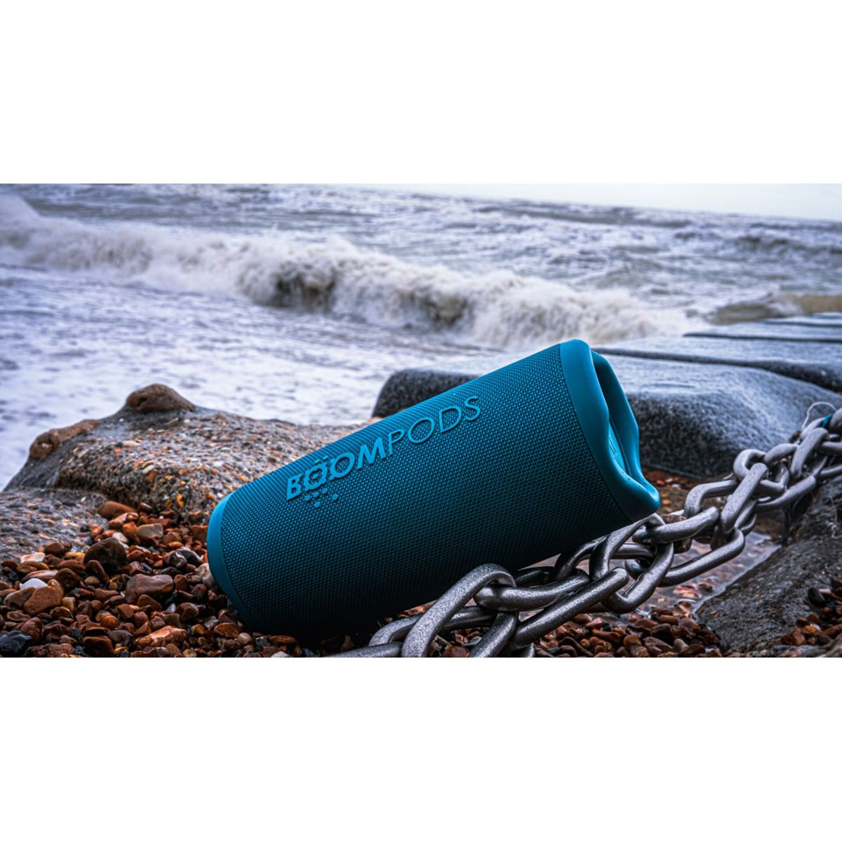 Altoparlante blu su spiaggia rocciosa, accanto a una catena, con onde sullo sfondo.